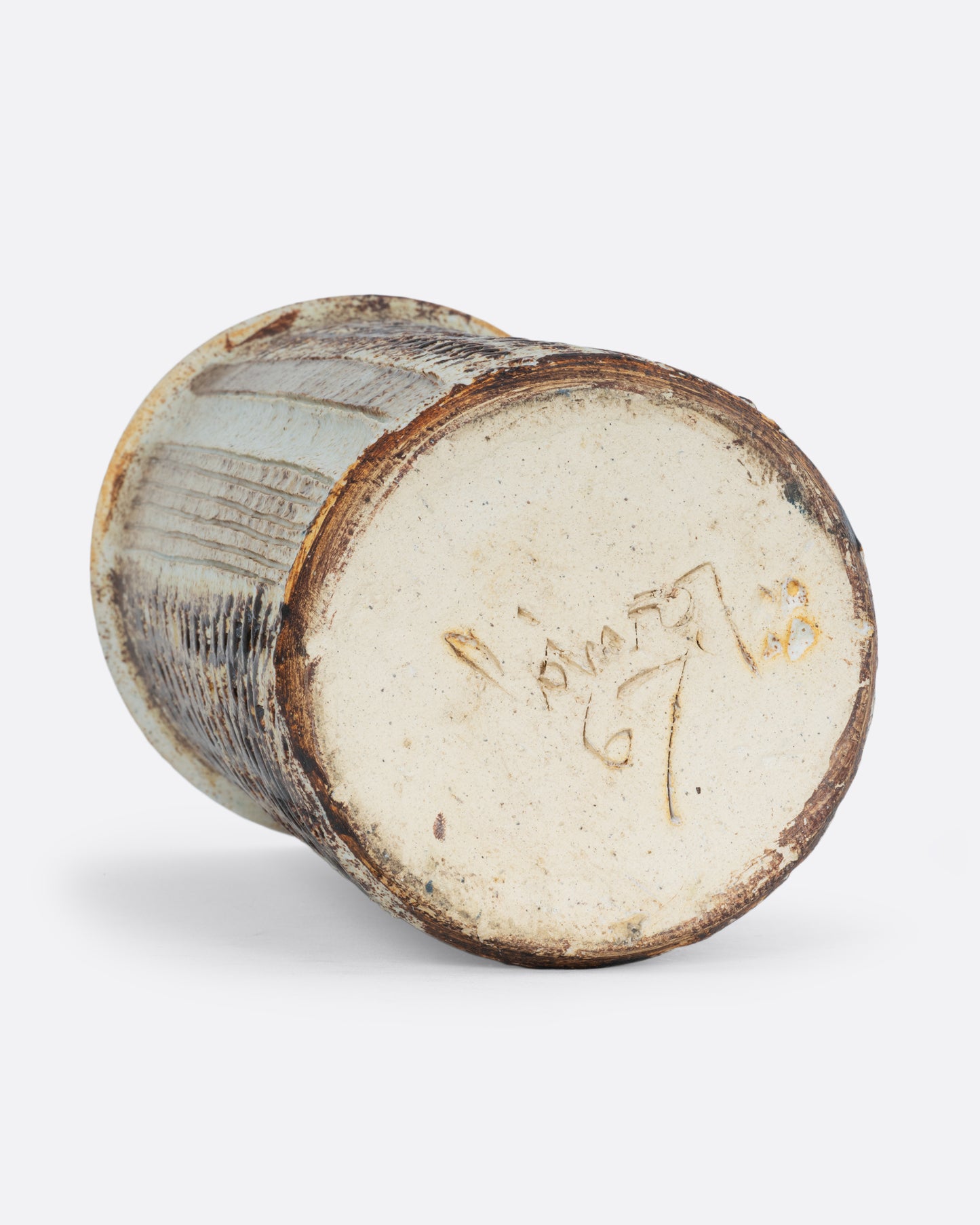 A tall textured tan vase with the artist signature and 67 on the bottom. Shown on its side from the bottom. 