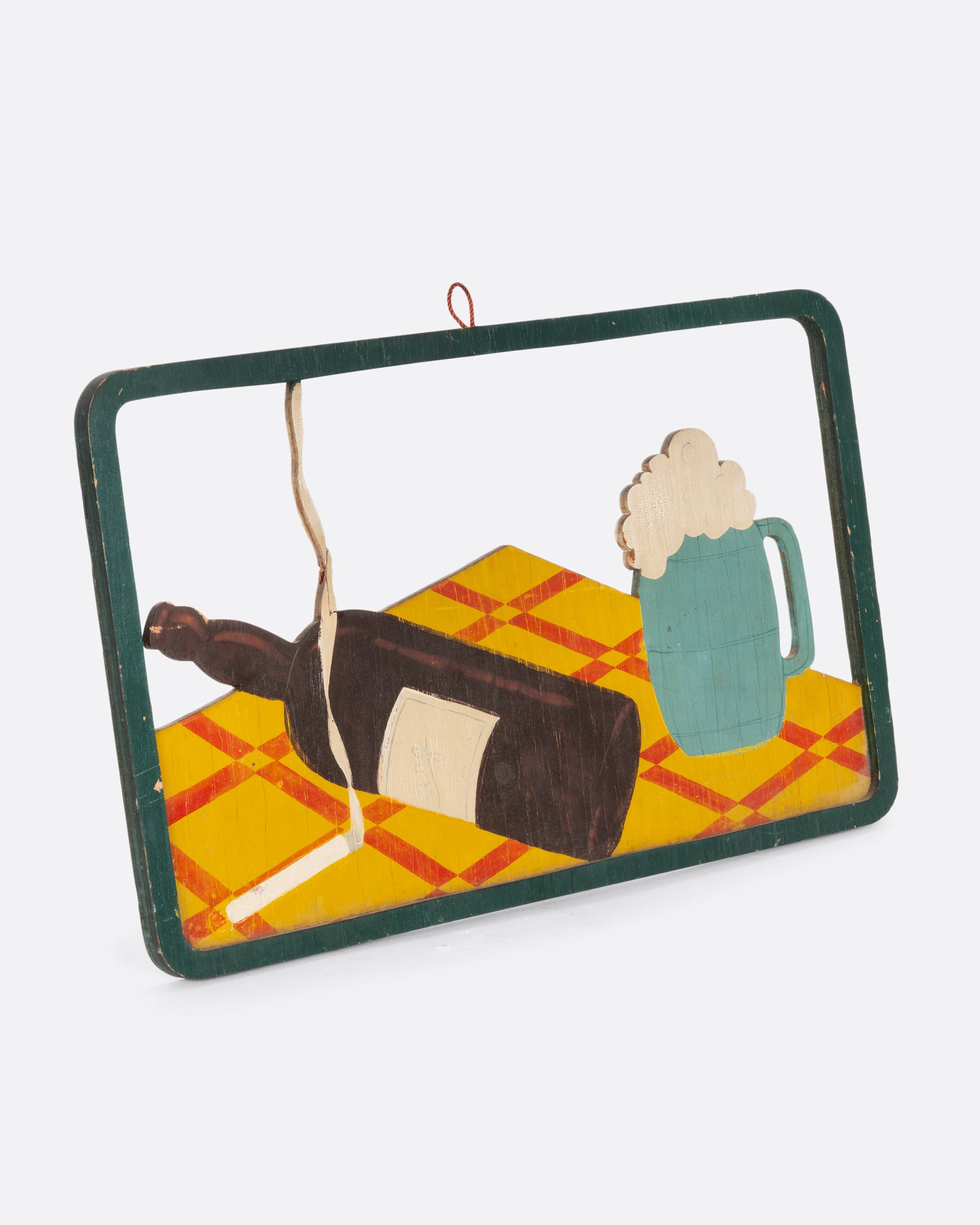 A framed wooden relief still life with a lit cigarette, wine bottle, and beer mug on a table top. Shown from the side.