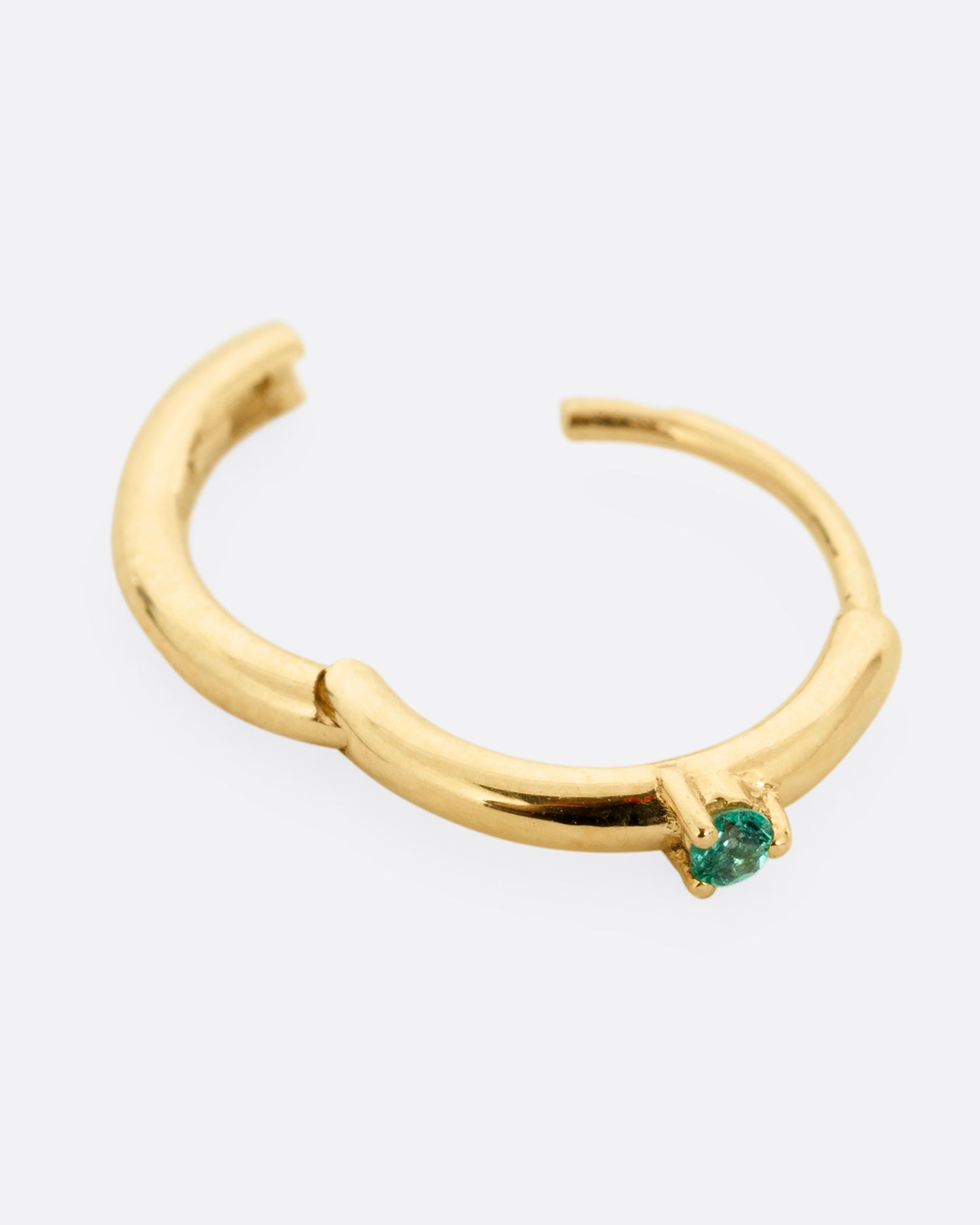 A yellow gold hinged huggie hoop with a prong set emerald at the front center. Shown open lying flat at an angle from the front.