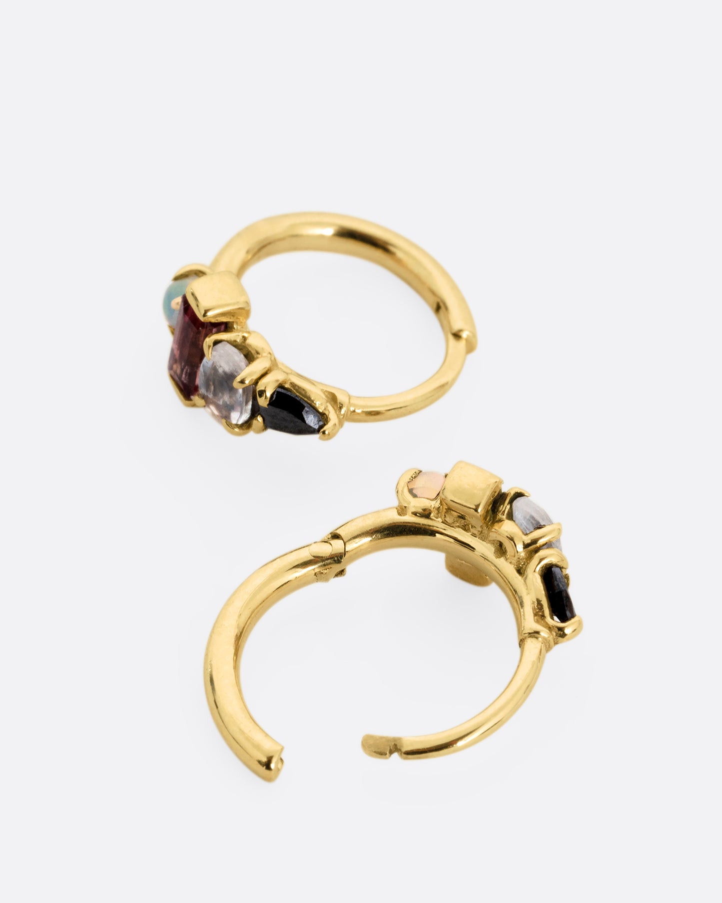 A pair of yellow gold huggie hoops with a gemstone stack of black diamond, moonstone, tourmaline, and opal. One earring is shown lying flat from the front, the other open from the back.