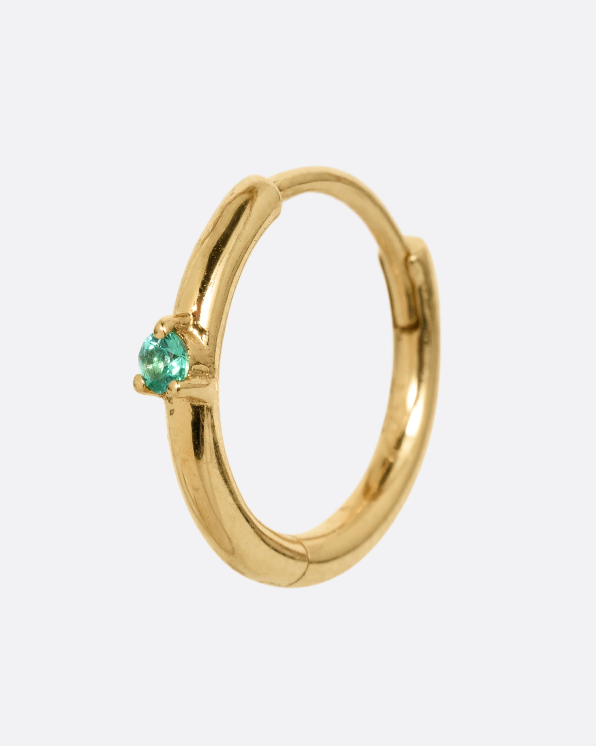 A yellow gold hinged huggie hoop with a prong set emerald at the front center. Shown at an angle from the front.