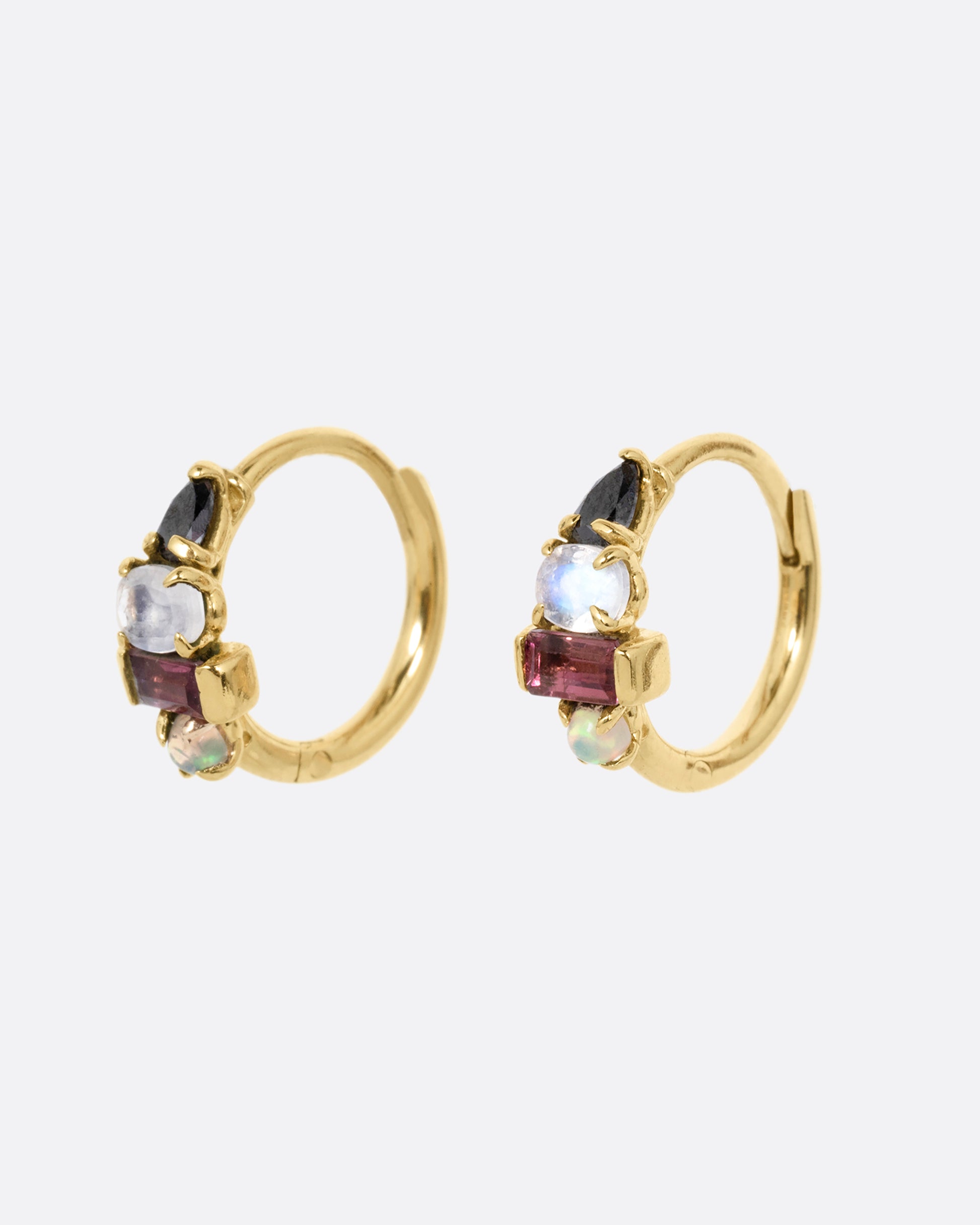 A pair of yellow gold huggie hoops with a gemstone stack of black diamond, moonstone, tourmaline, and opal. Shown from the side.