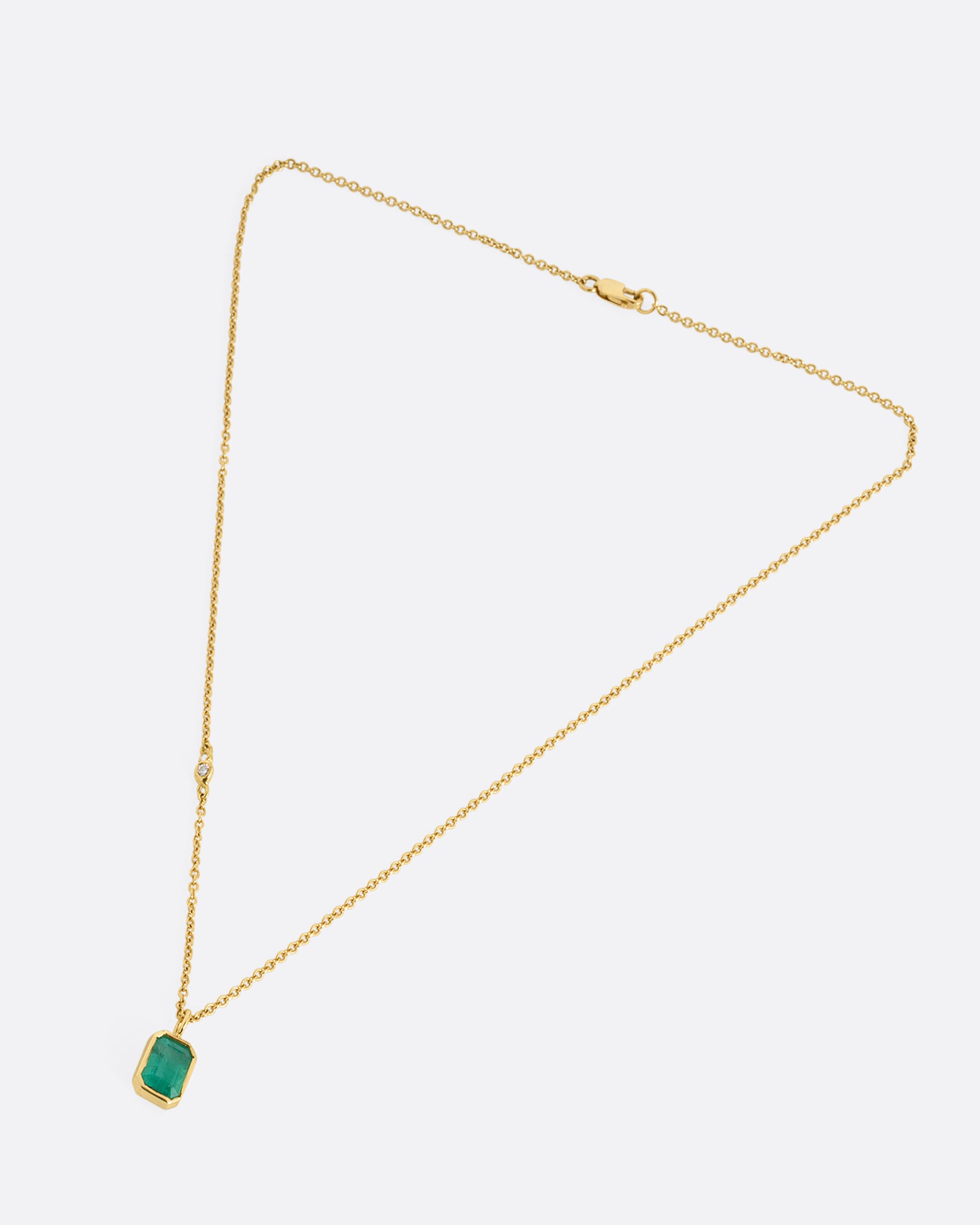 A yellow gold cable chain necklace with a bezel set Zambian emerald and diamond details on one side. Shown closed lying flat from the front.
