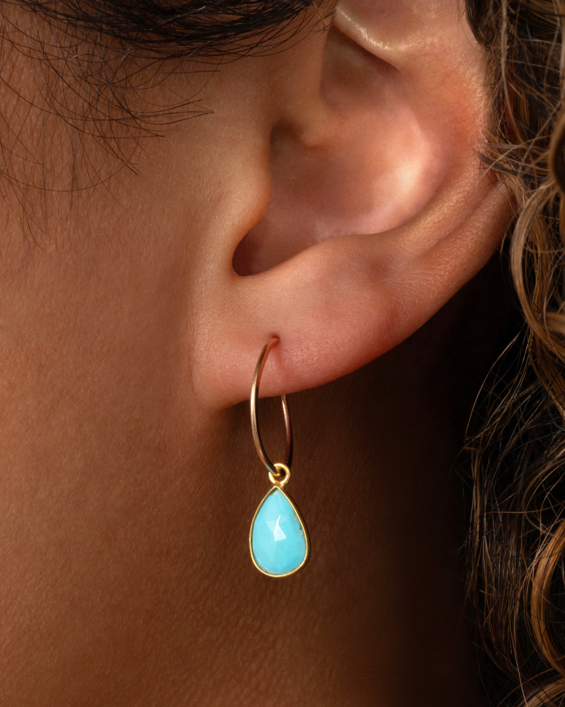 A turquoise teardrop shaped dangle set in yellow gold with a yellow gold bail. Shown from the front on a yellow gold hoop on a person.