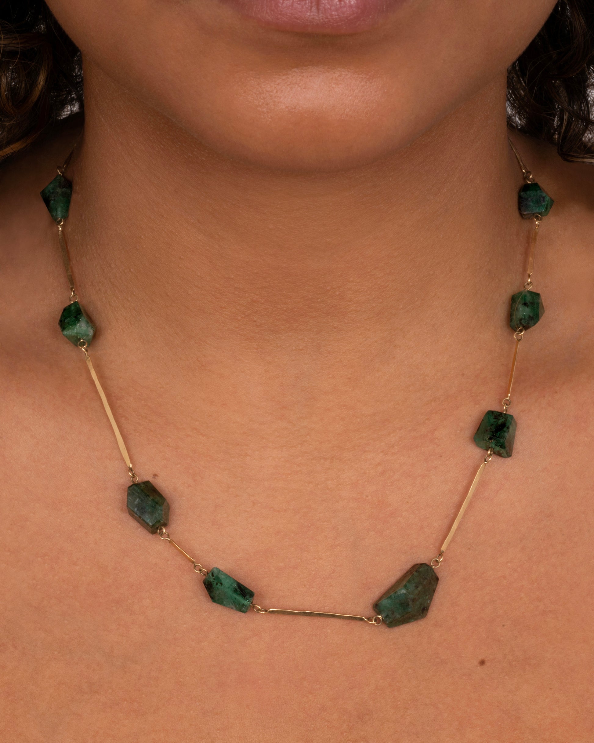 A yellow gold hammered bar link necklace with raw emeralds throughout. Shown worn around a neck.