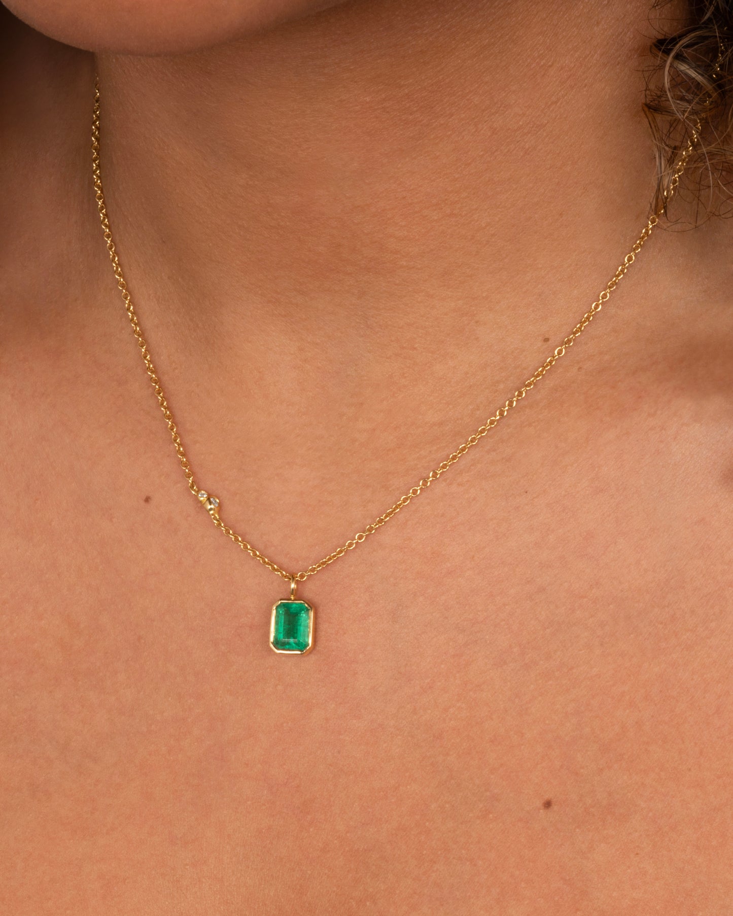 A yellow gold cable chain necklace with a bezel set Zambian emerald and diamond details on one side. Shown from the front on a person.