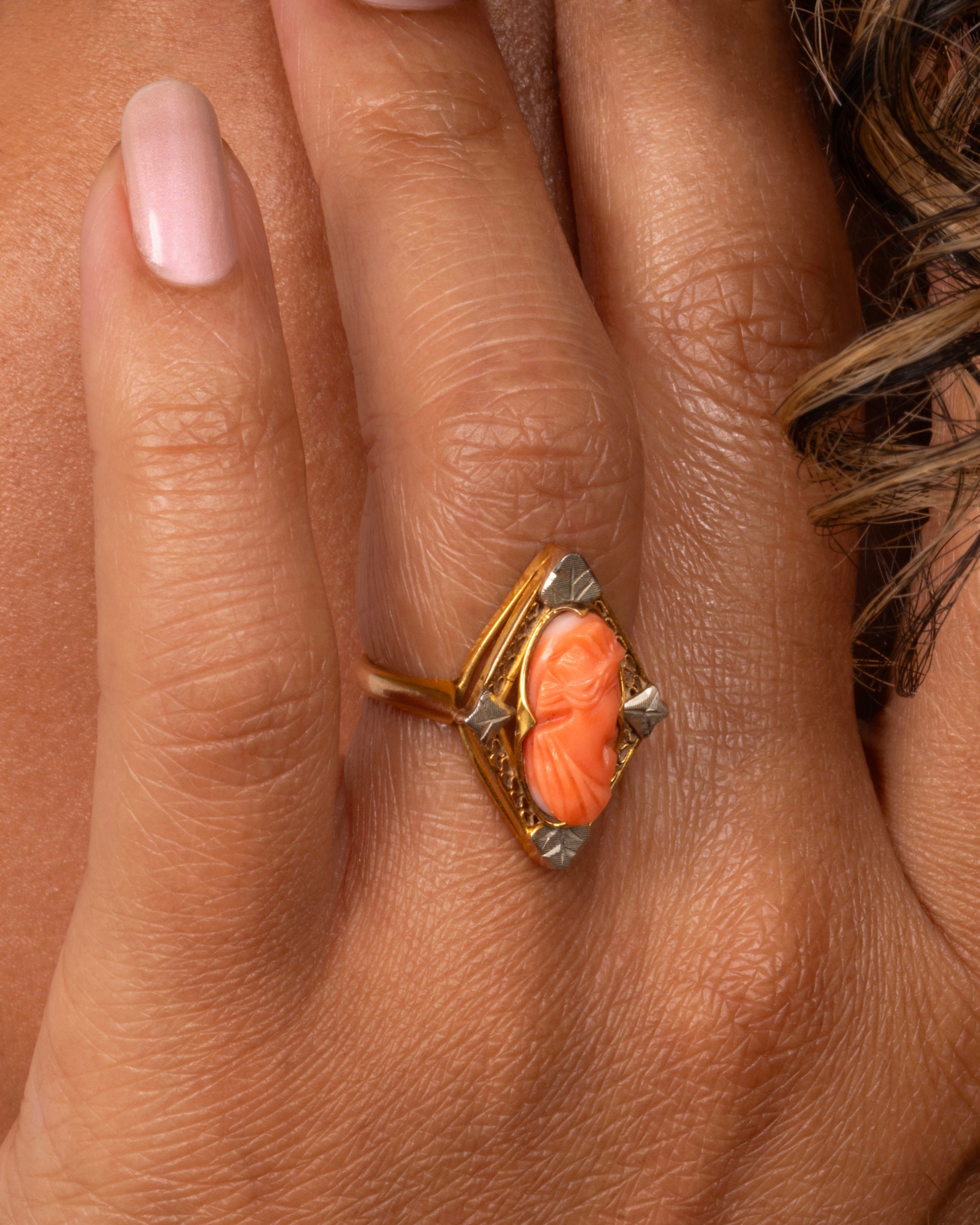 A yellow gold cameo ring with a coral carved portrait. Shown on a hand.
