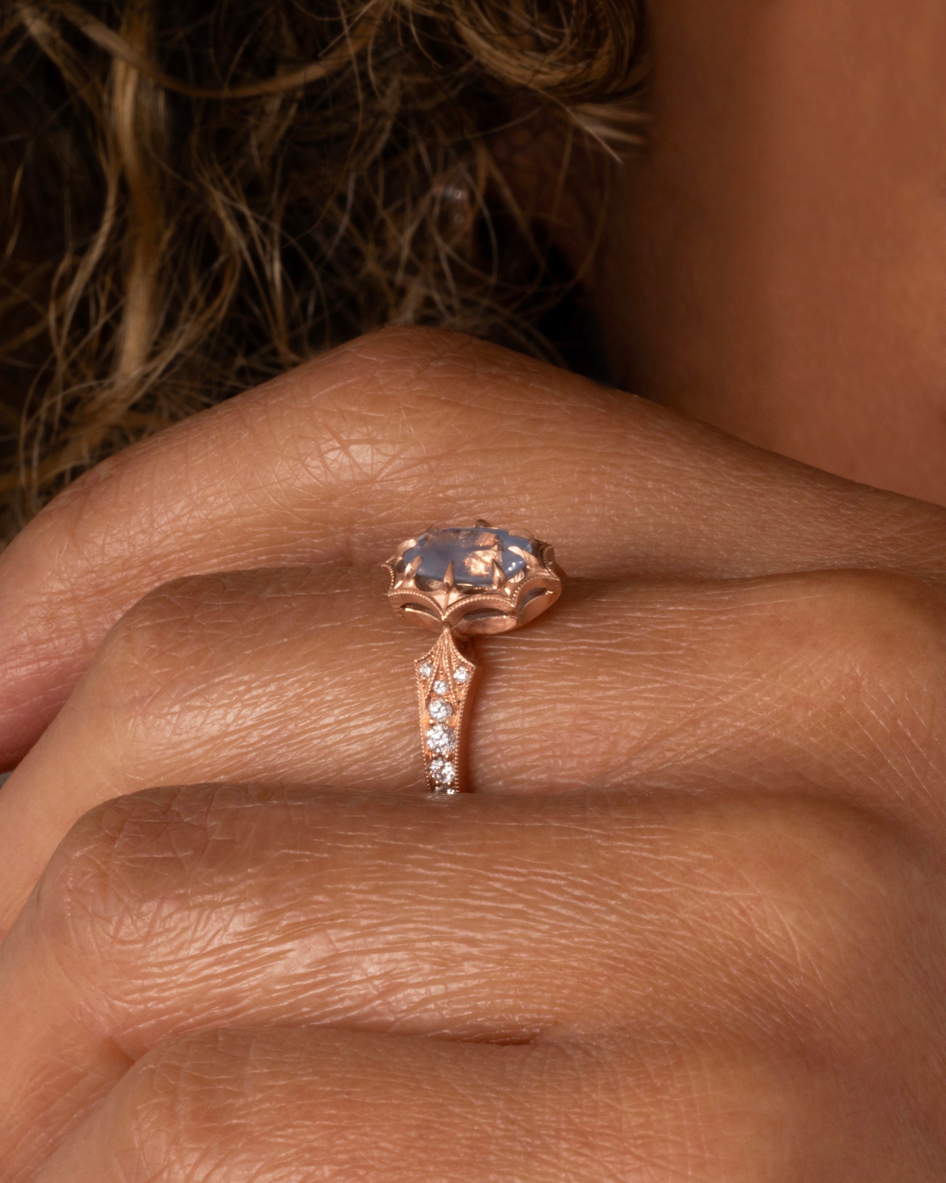 A rose gold ring with a prong set lavender sapphire at the center and diamond lined shoulders. Shown worn on a hand.