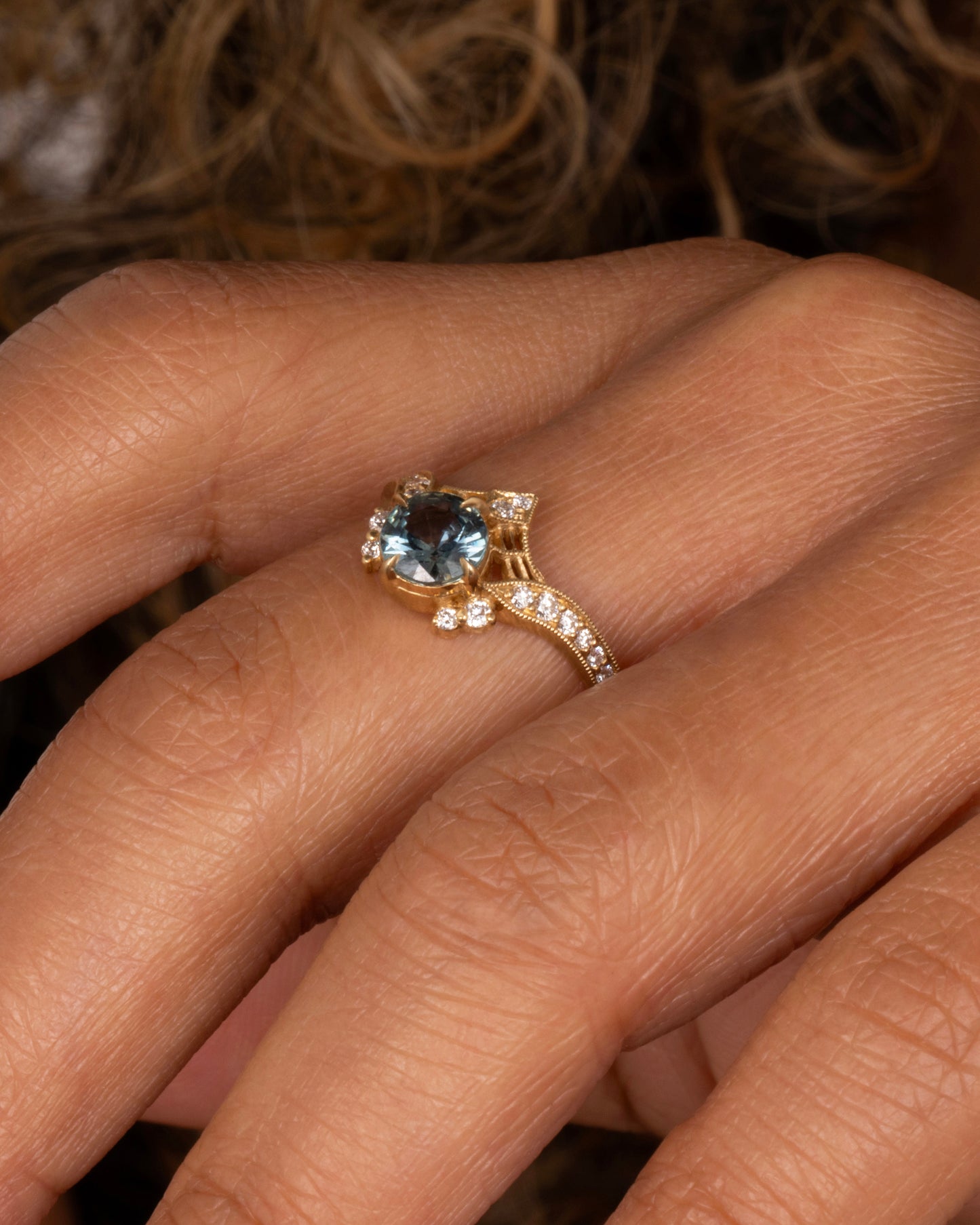 A yellow gold ring with a prong set teal sapphire, delicate cutouts, and white diamond accents. Shown on a finger.