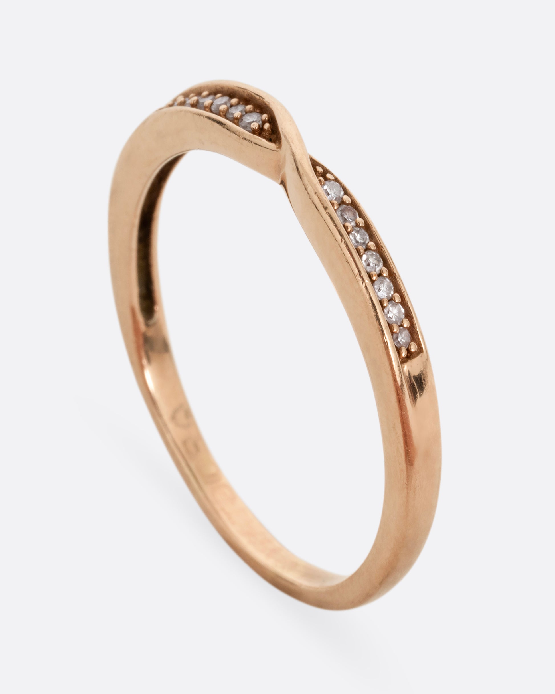 A rose gold ring with a twist at the center and seven white diamonds on either side. Shown standing up from the side.