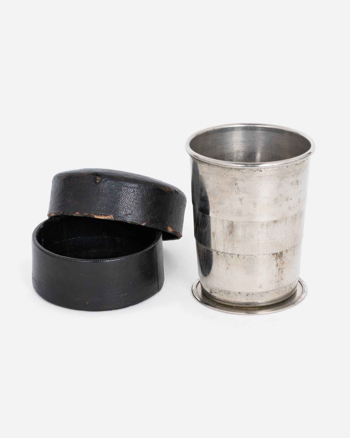 A silver  collapsible cup with a round leather case open to its side. Shown from the front.
