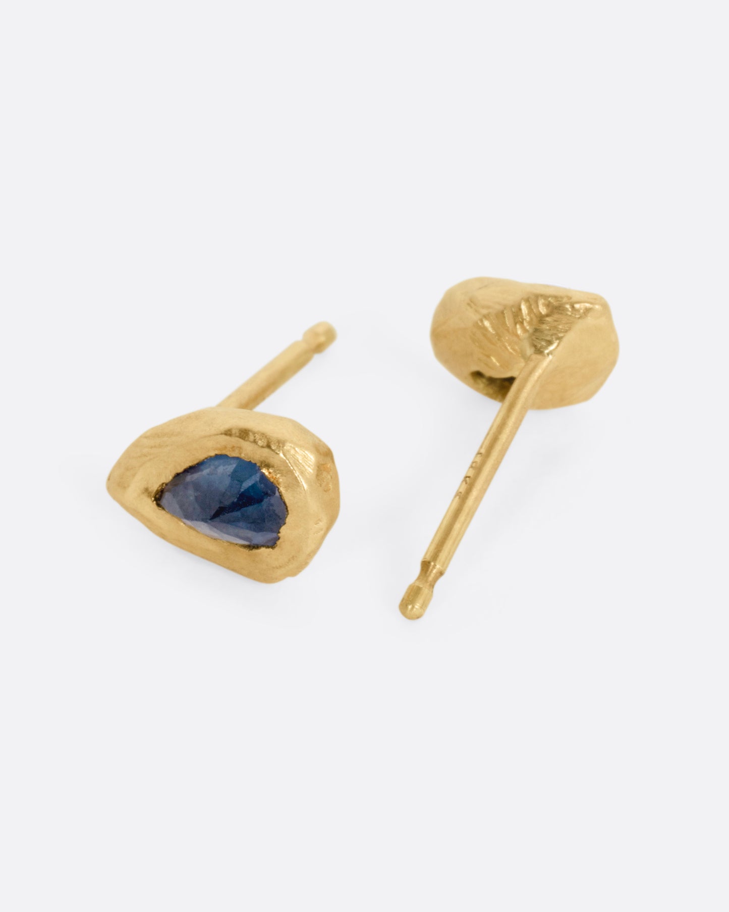 A pair of yellow gold stud earrings with teardrop shaped blue sapphires, shown on their sides. One earring is facing front, the other, the back.