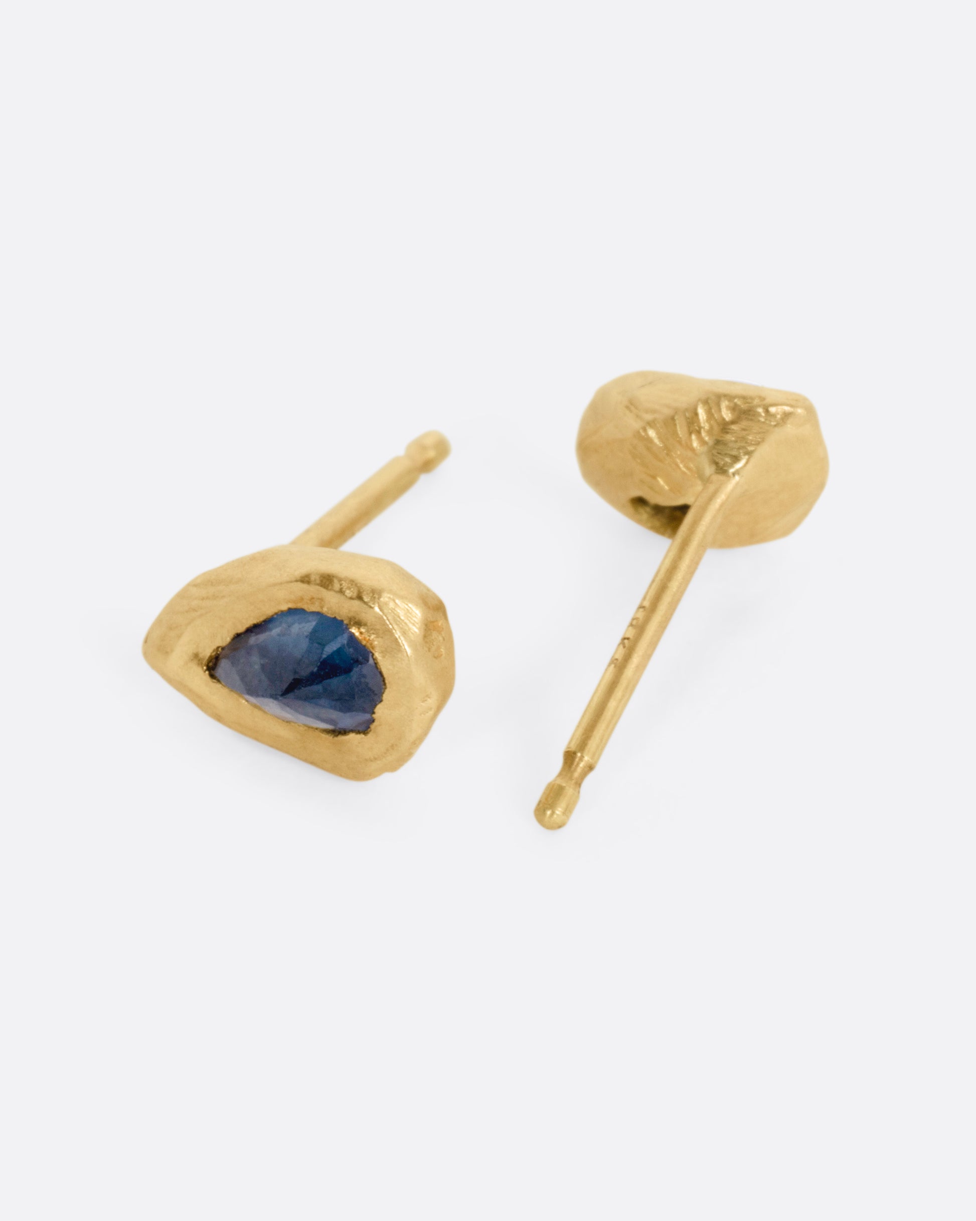 A pair of yellow gold stud earrings with teardrop shaped blue sapphires, shown on their sides. One earring is facing front, the other, the back.