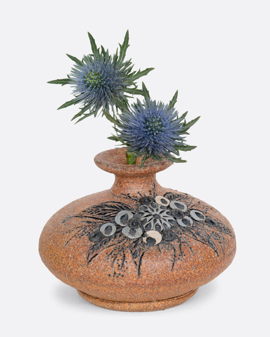 A brown stoneware pottery vase with a floral motif on the side. Shown with blue flowers.