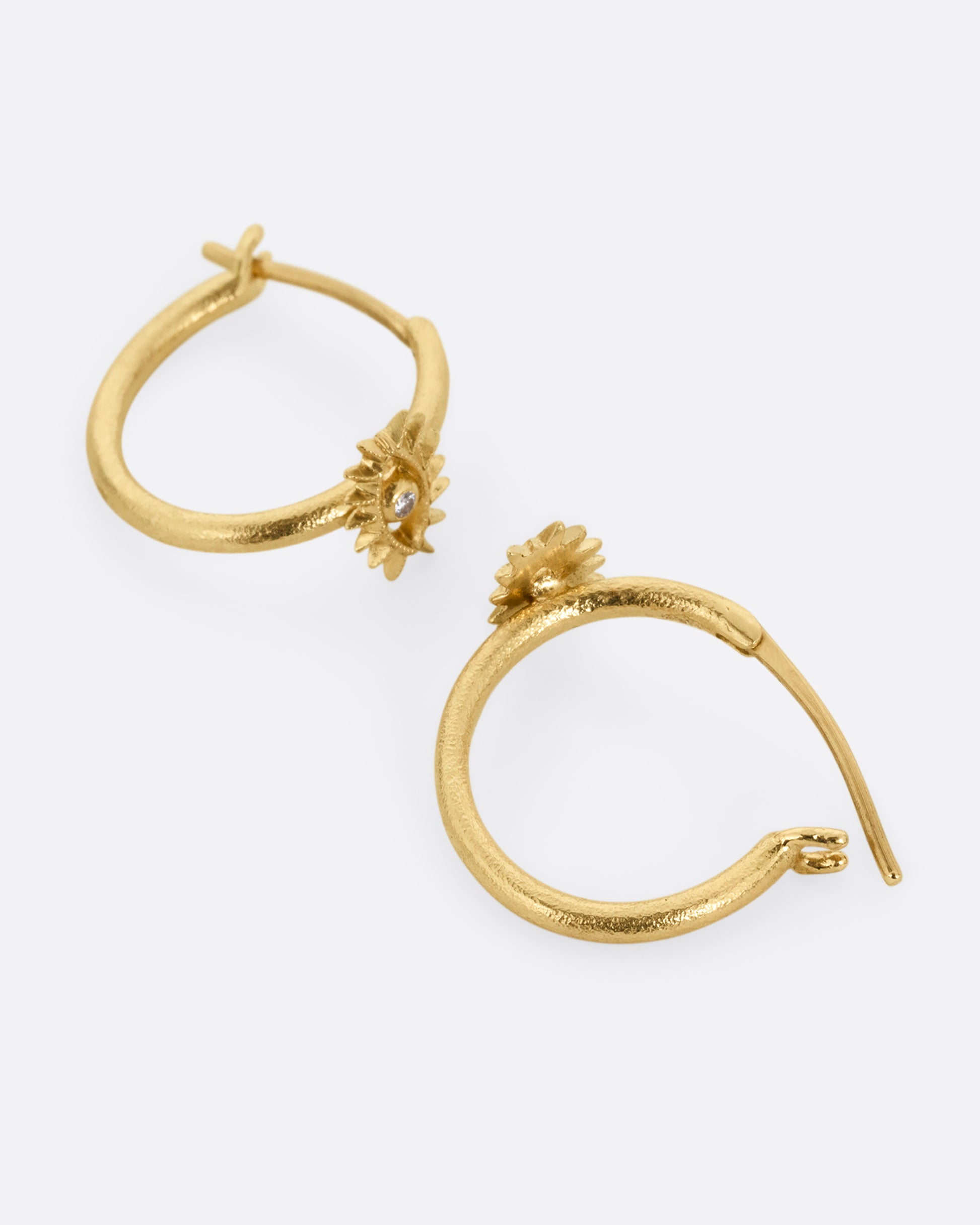 A pair of yellow gold hoop earrings with diamond studded eyes with dramatic lashes. Shown lying on their side, one from the front, the other open from the back.