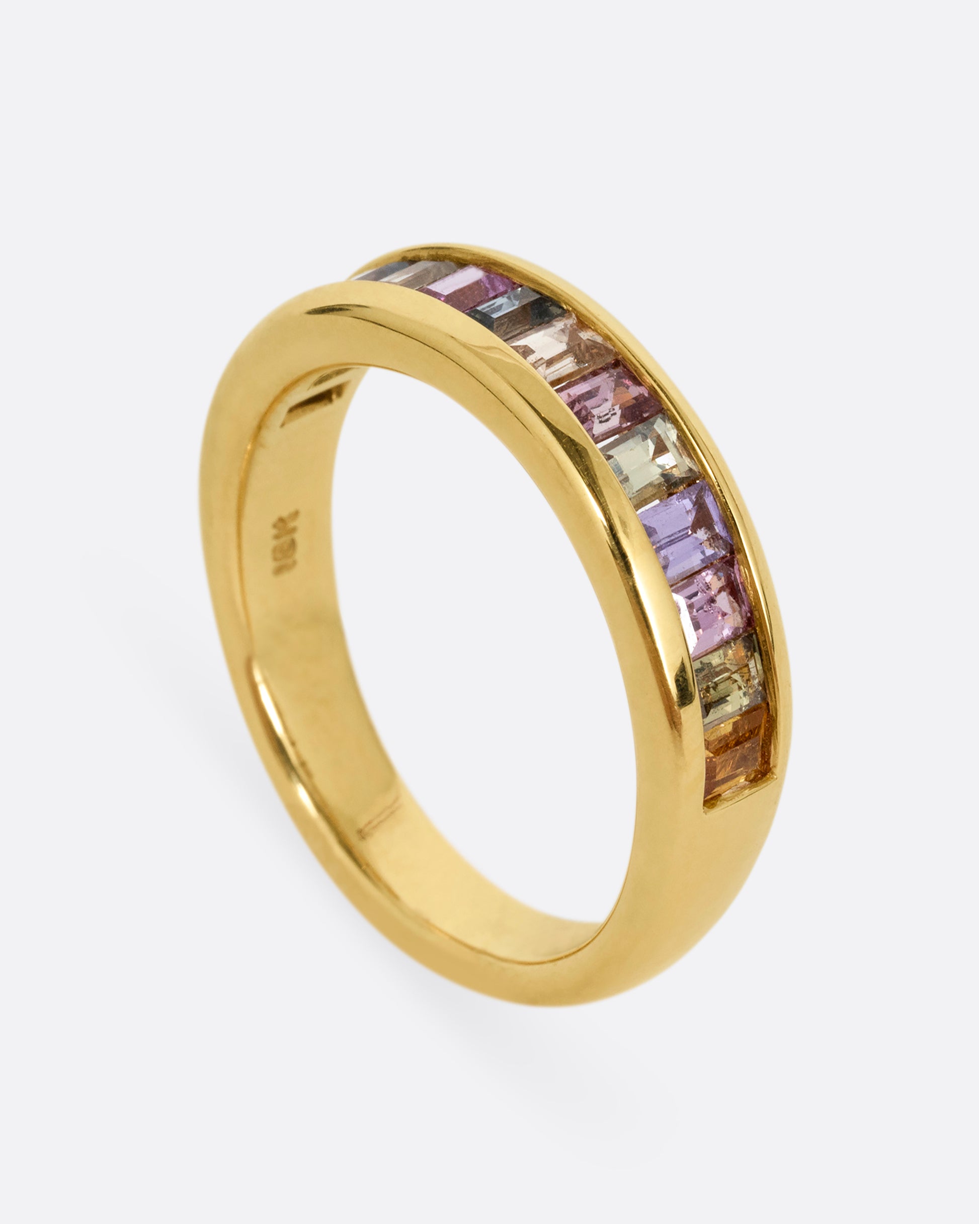 A yellow gold ring with channel set multicolored sapphires. Shown standing up from the side.