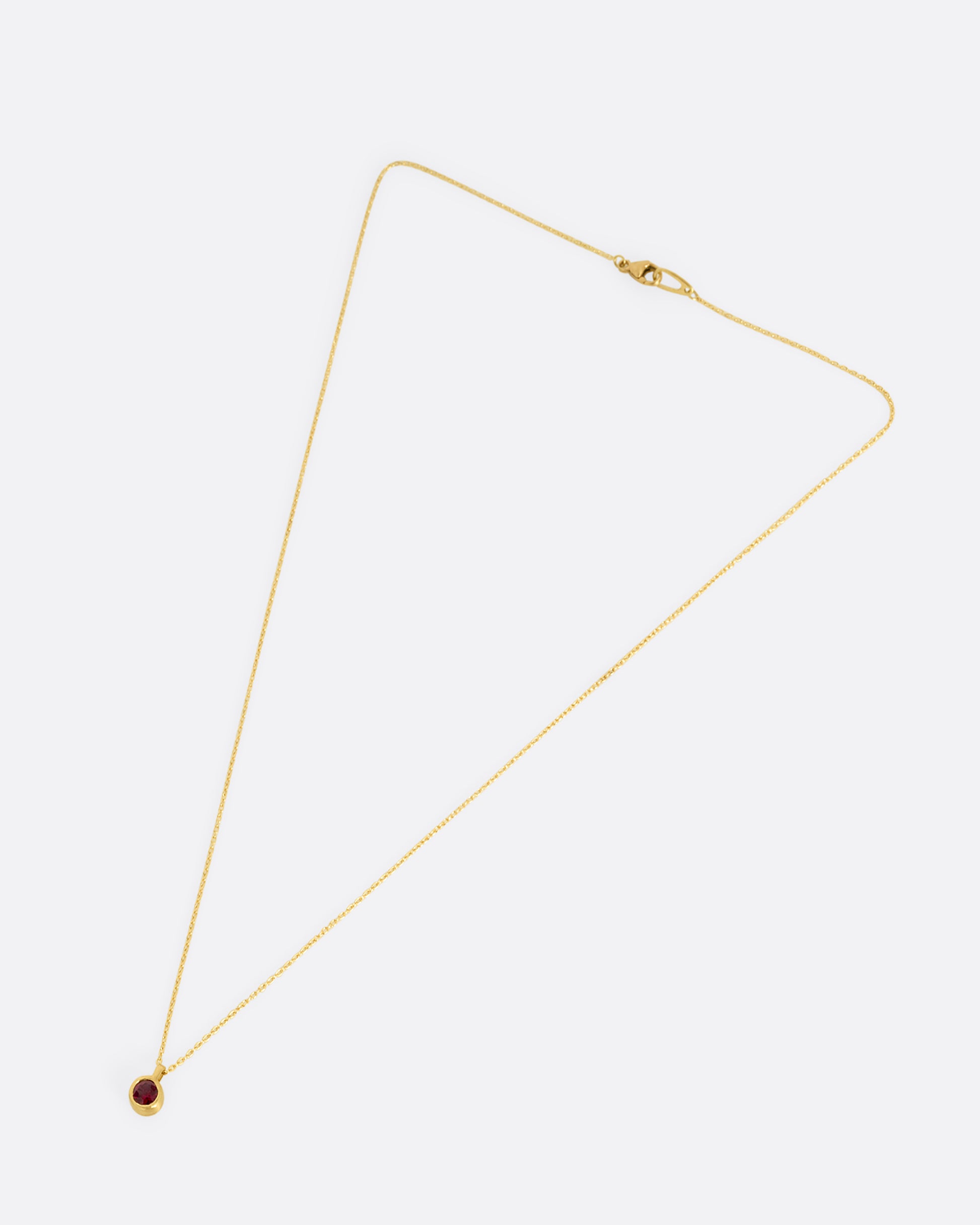 A yellow gold cable chain necklace with a single round ruby in a brushed gold bezel. Shown clasped from the front lying flat.