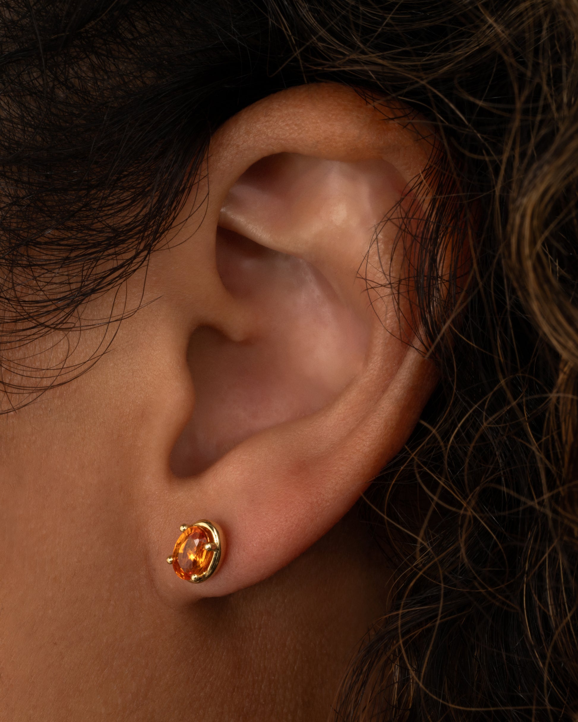 A yellow gold stud earring with an orange sapphire. Shown from the front on a person.