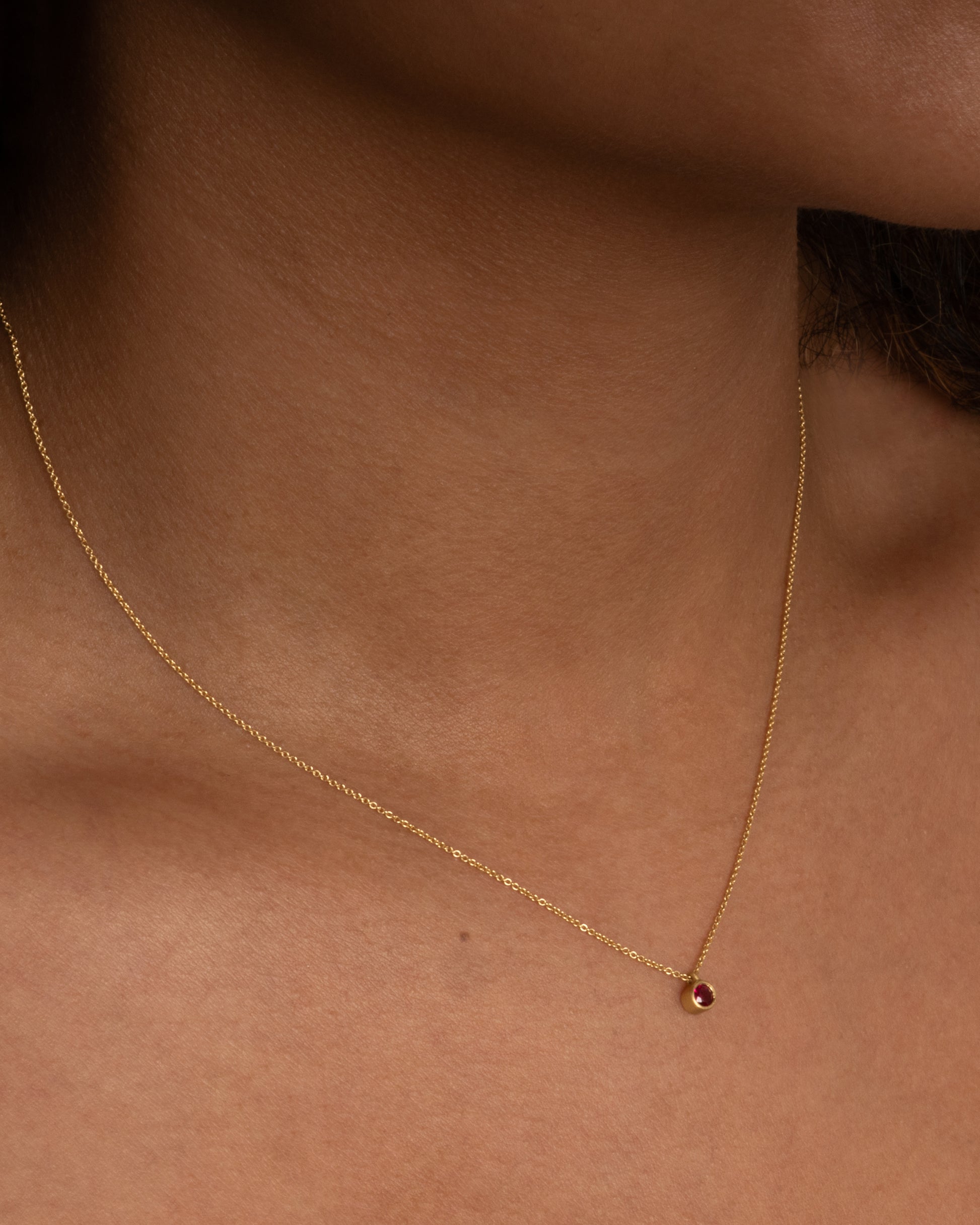 A yellow gold cable chain necklace with a single round ruby in a brushed gold bezel. Shown from the front on a person.