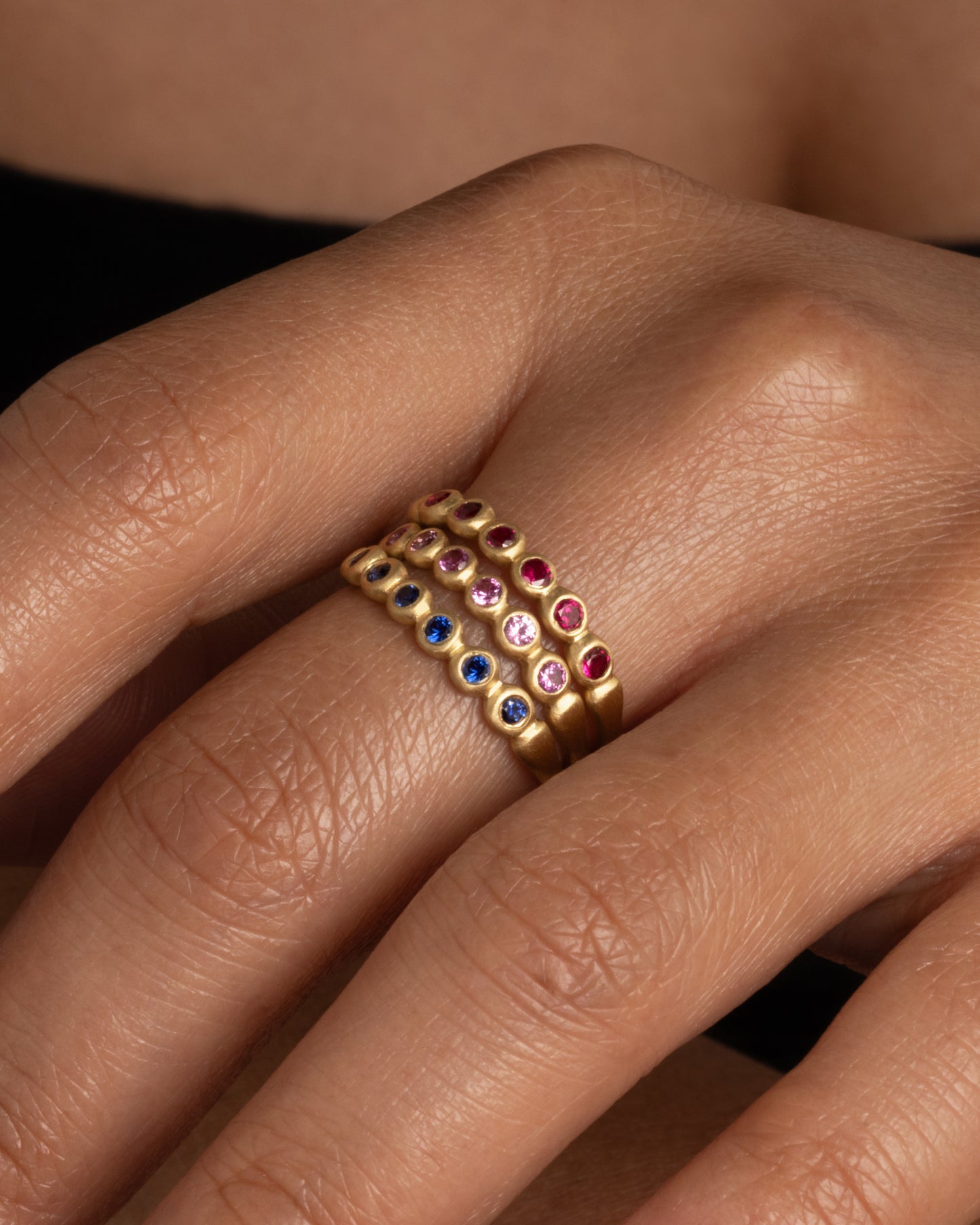 Three yellow gold rings with bezel set blue and pink sapphires and rubies. Shown stacked from the front on a hand.