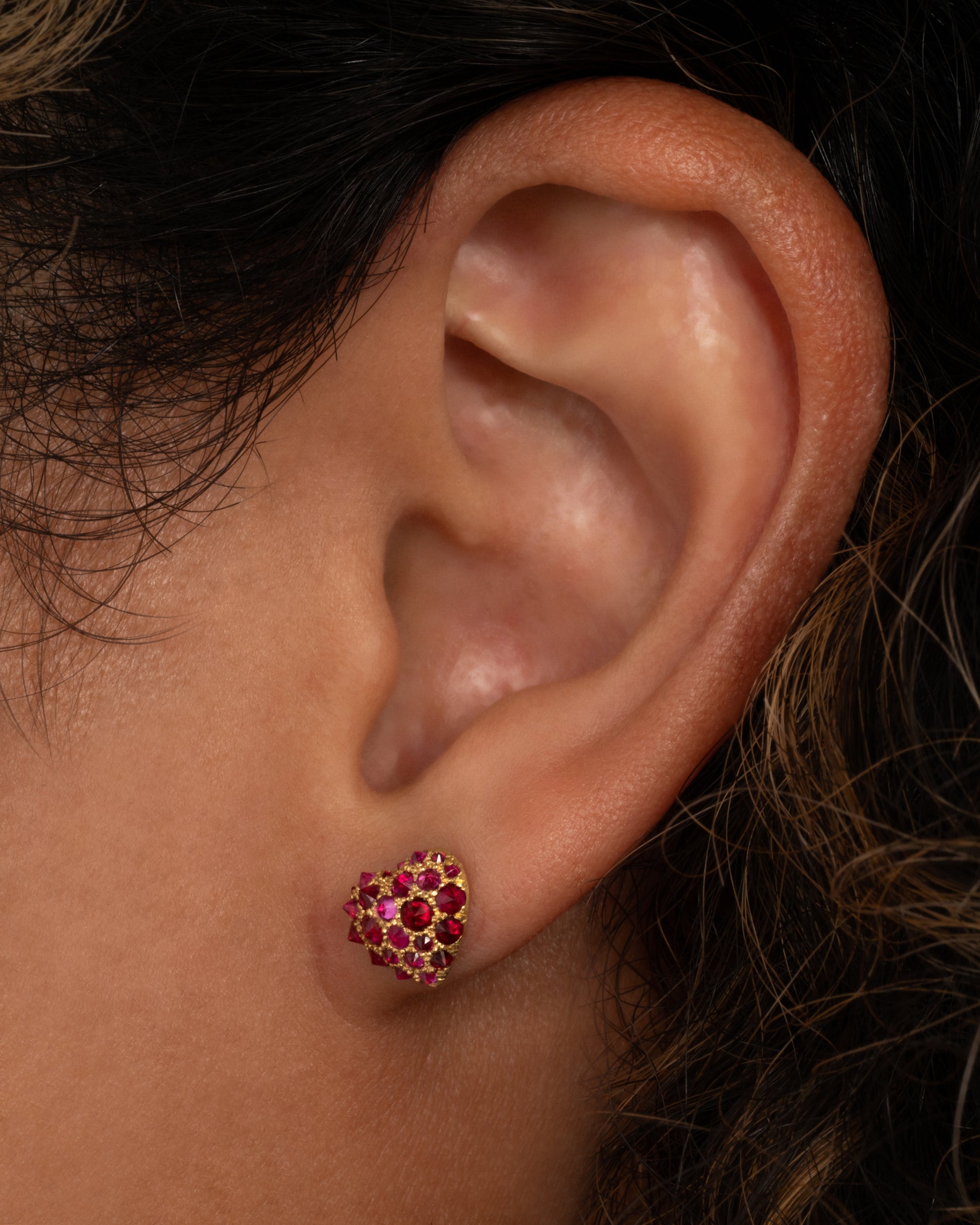 A yellow gold heart shaped stud earring with reverse set rubies of varying sizes set into them. Shown on an ear from the front.