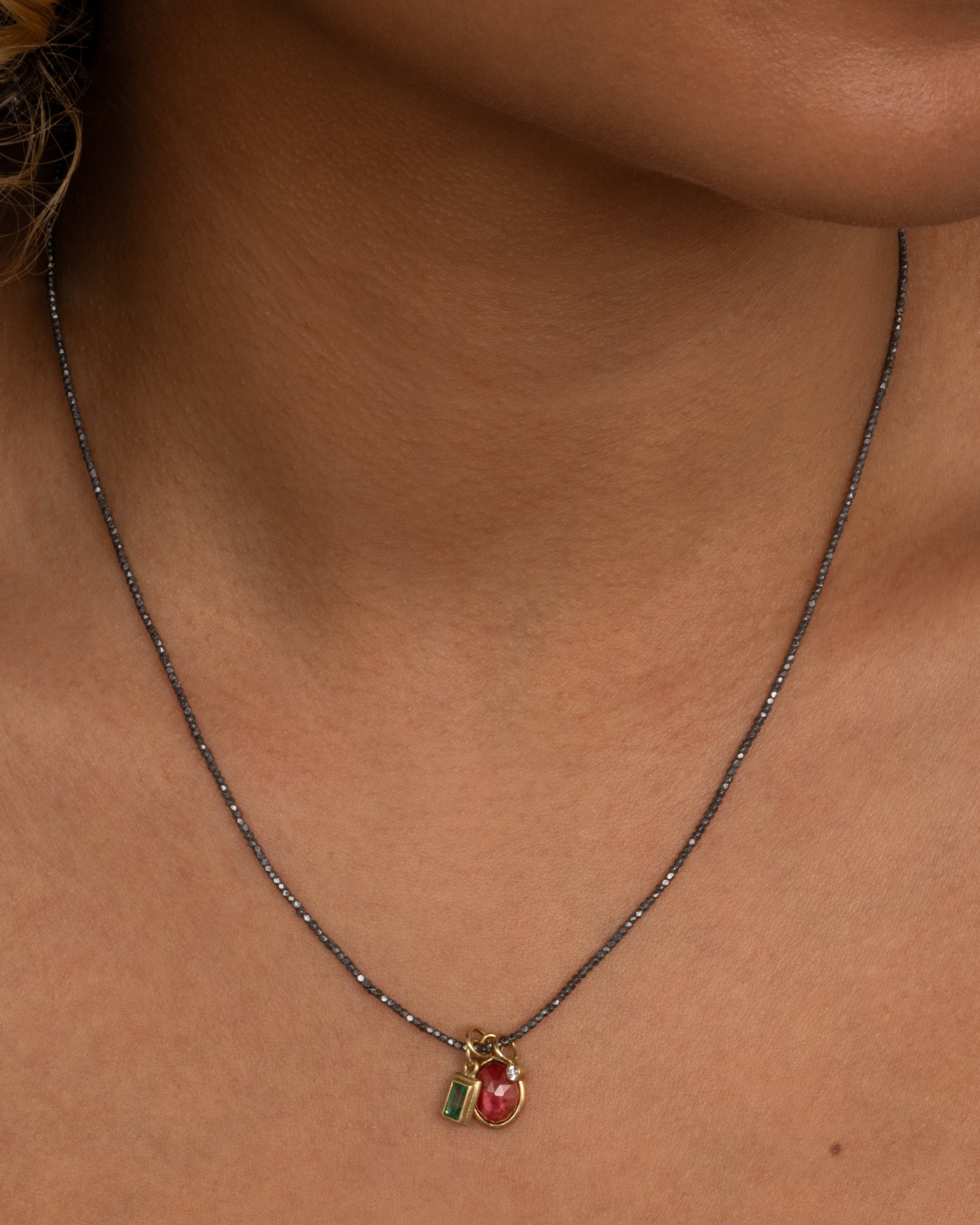 A necklace with three charms; pink tourmaline, emerald, and diamond with oxidized silver beads. Shown from the front on a person.