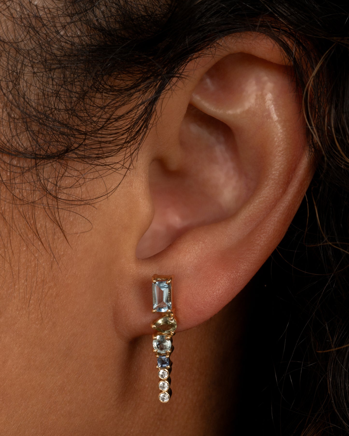 A yellow gold stud earring with a mosaic of aquamarine, tanzanite, apatite and diamonds. Shown worn in an ear.