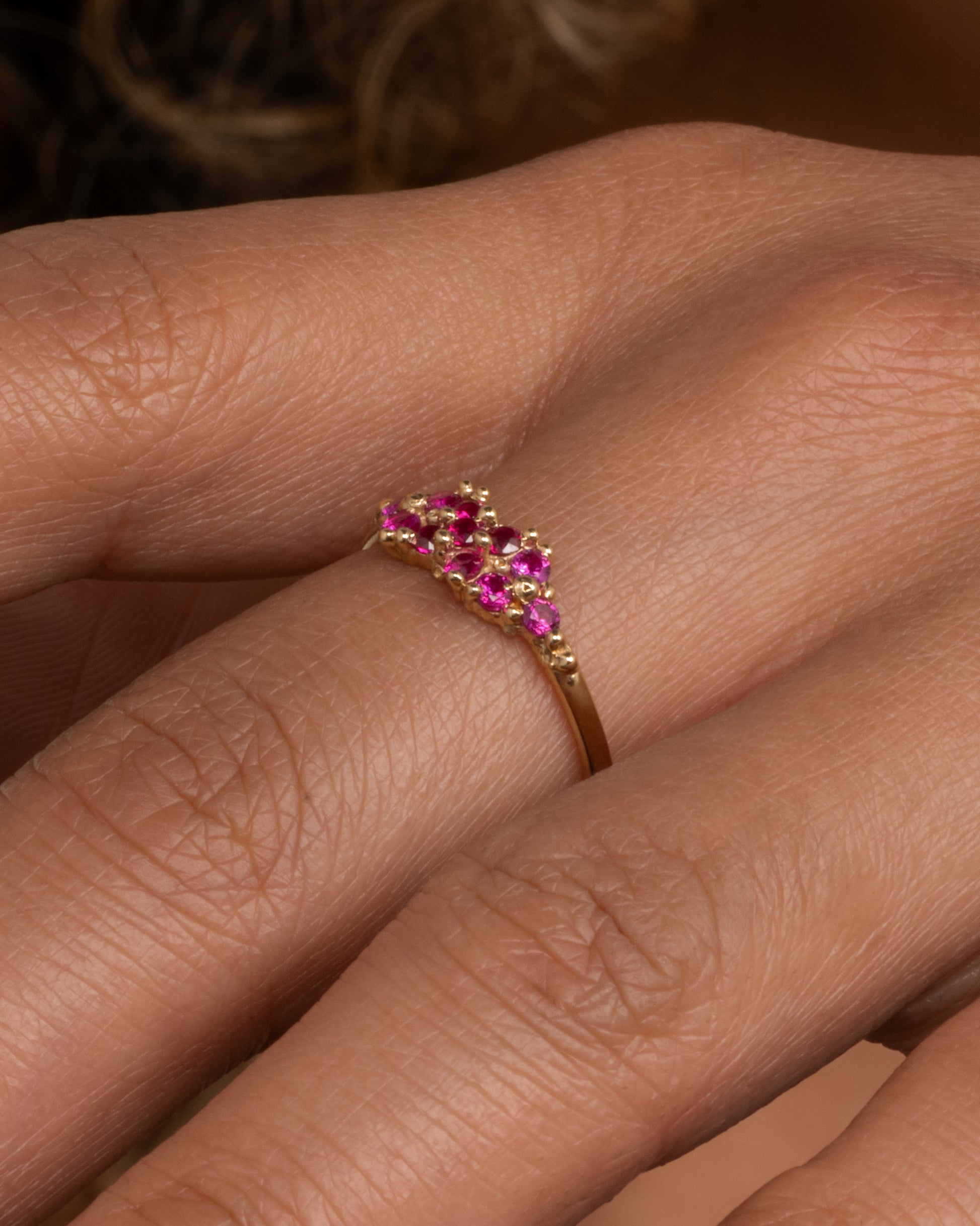 A yellow gold ring with a cluster of rubies arranged on the face. Shown worn on a hand.