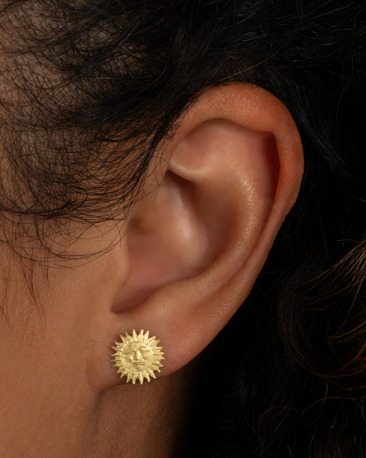A yellow gold sun stud earring with a face. Shown worn in an ear.