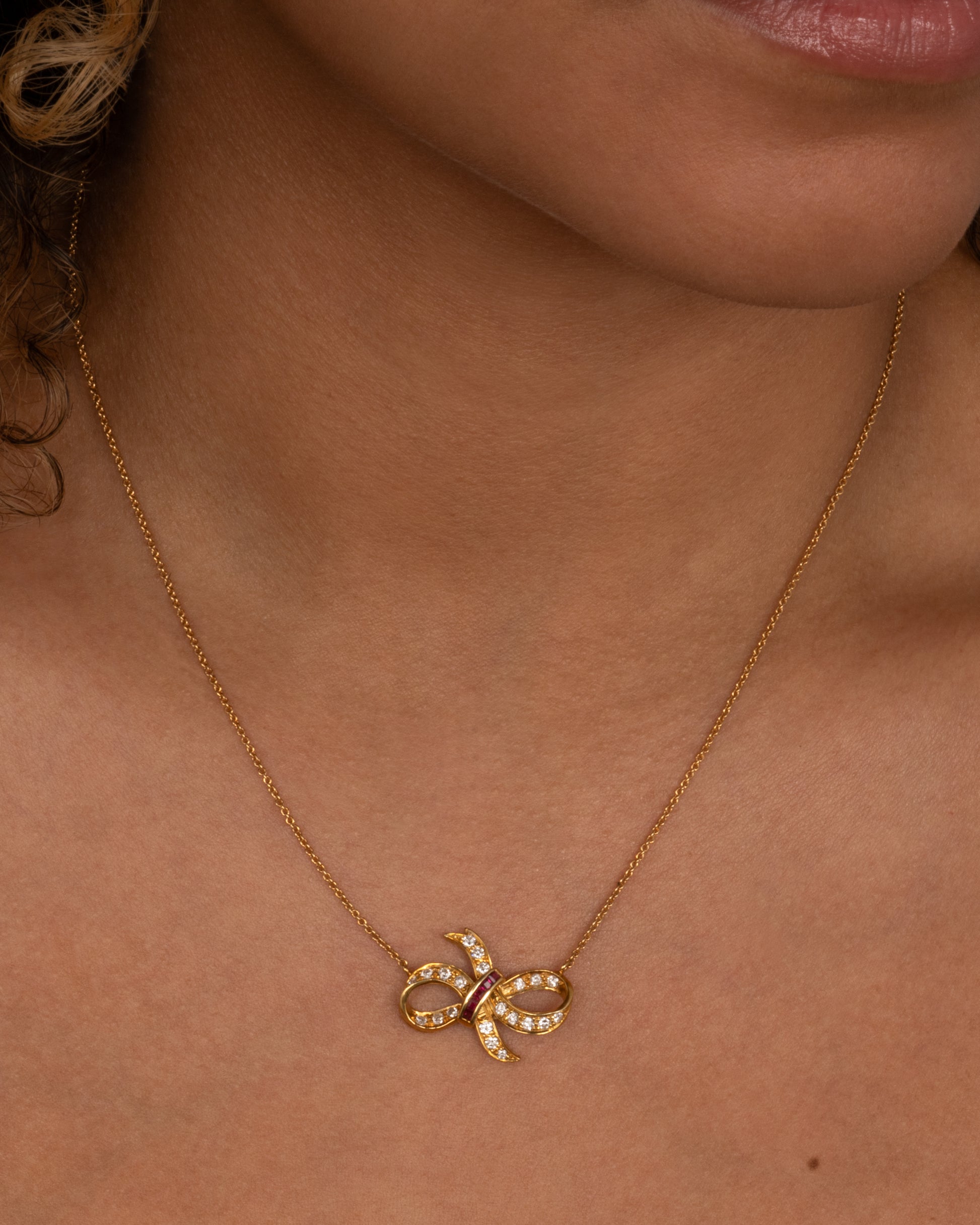 A yellow gold cable chain necklace with a diamond and ruby studded bow at the center. Shown from the front on a person.