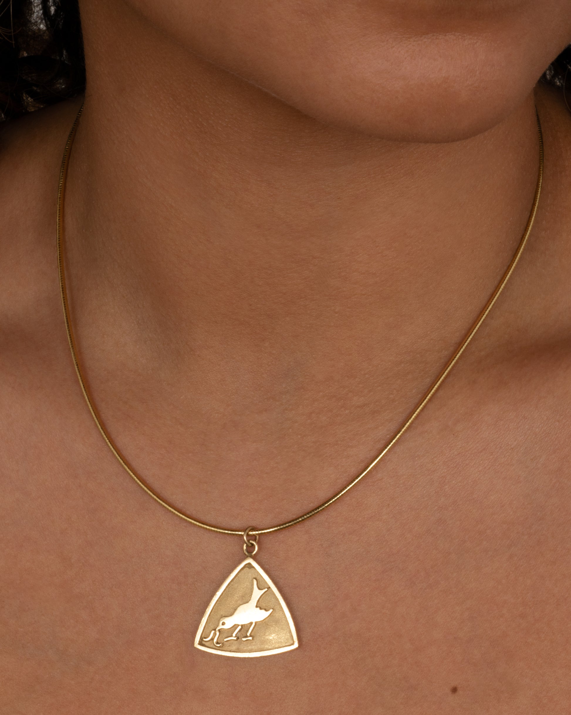 A yellow gold triangular charm with a bird getting worm on the front. Shown styled on a chain worn around the neck.