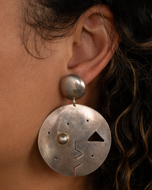A round drop stud earring with geometric cutout face. Shown from the  front on a person.