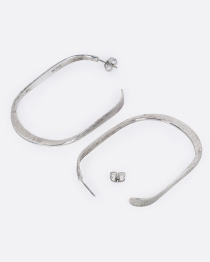A pair of oval silver hammered hoop earrings, shown from the side with one hoop open.