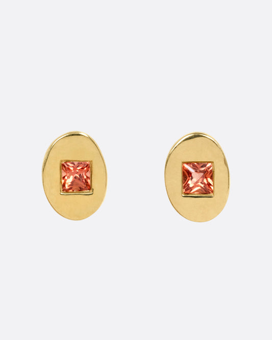 A pair of yellow gold oval stud earrings with princess cut orange sapphires, shown from the front.