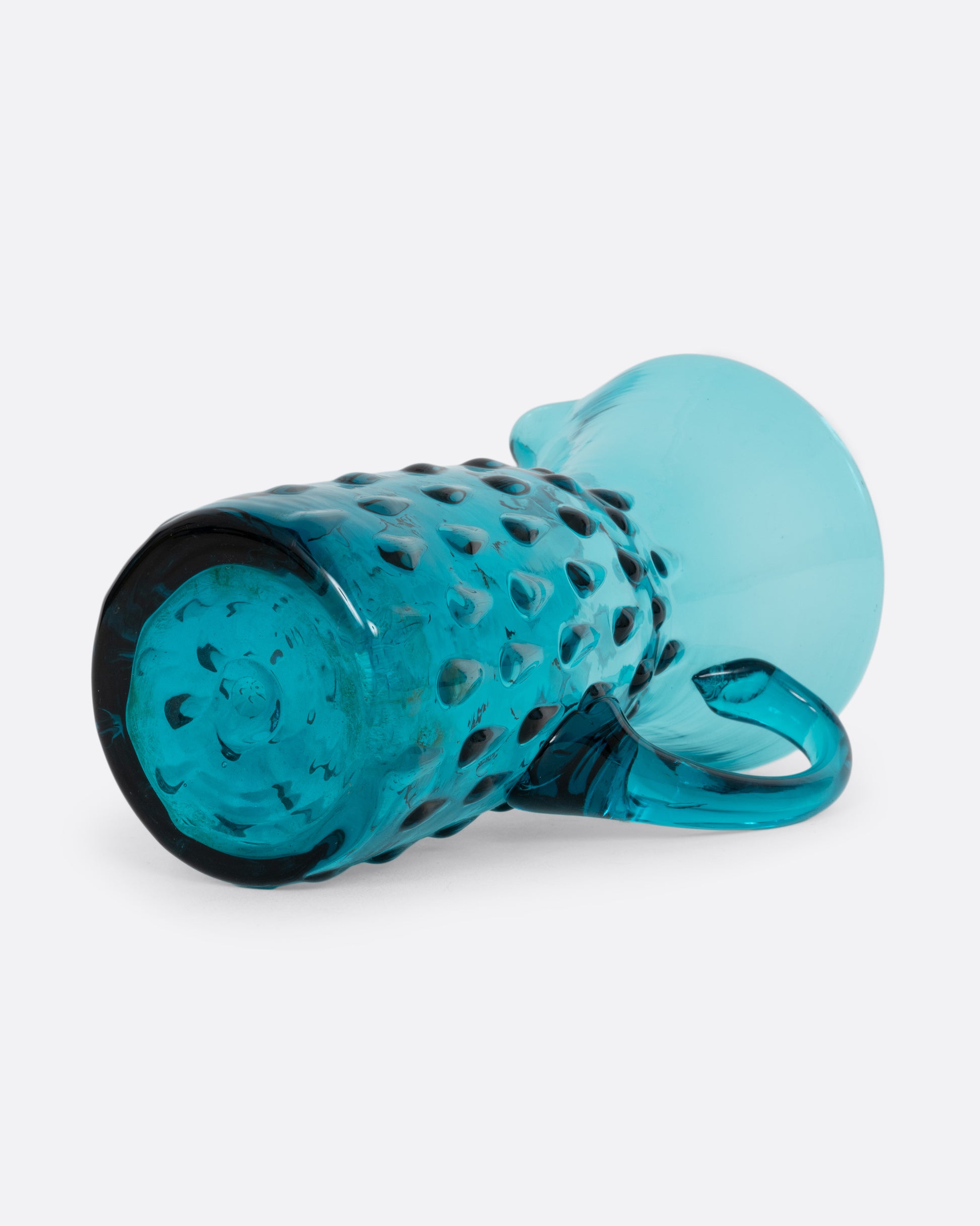 A teal glass creamer with a funnel shaped top, handle, and a bubbly teardrop pattern.  Shown lying on its side from the bottom.