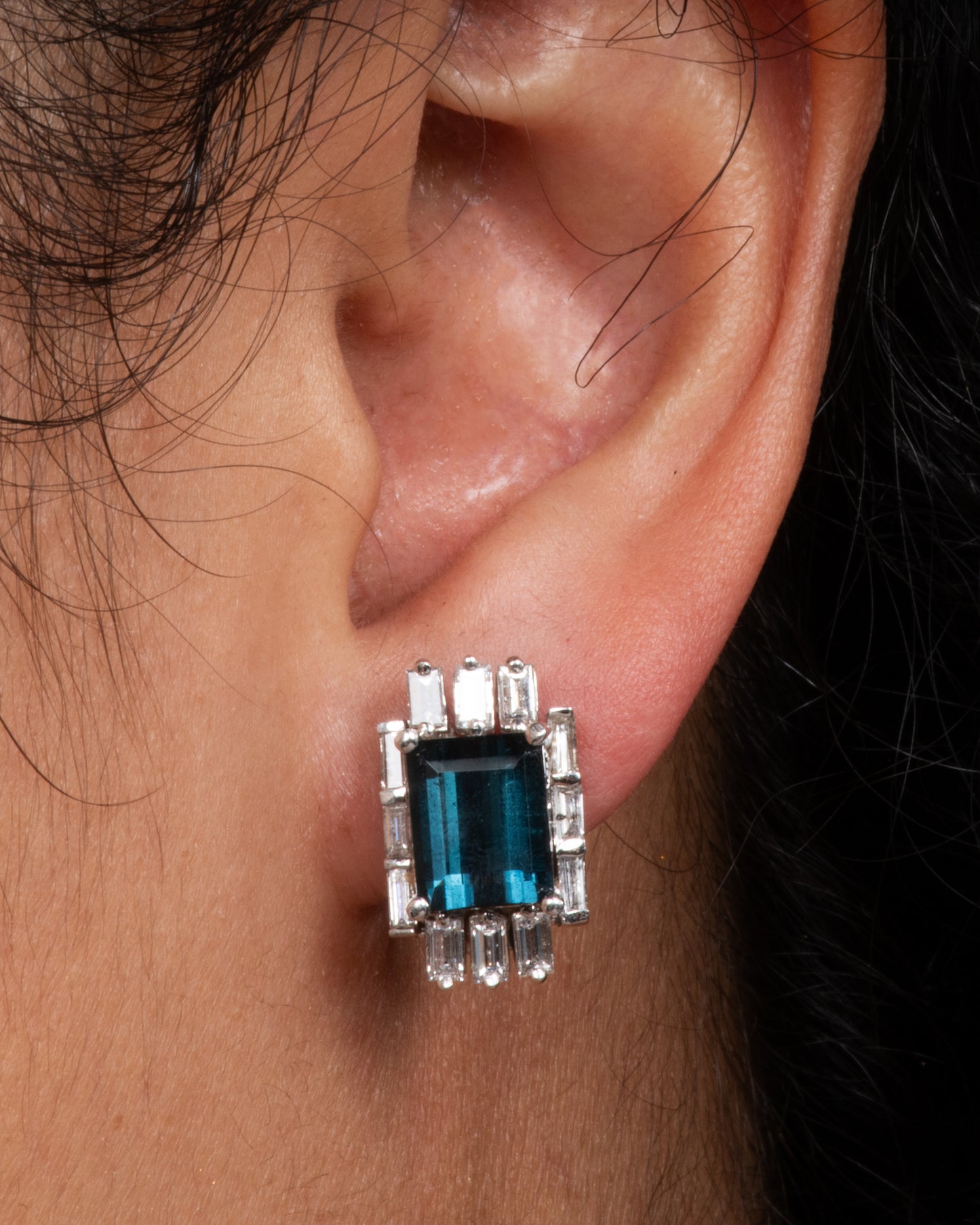 A pair of white gold earrings with emerald cut green tourmalines surrounded by baguette diamonds. View on an ear.