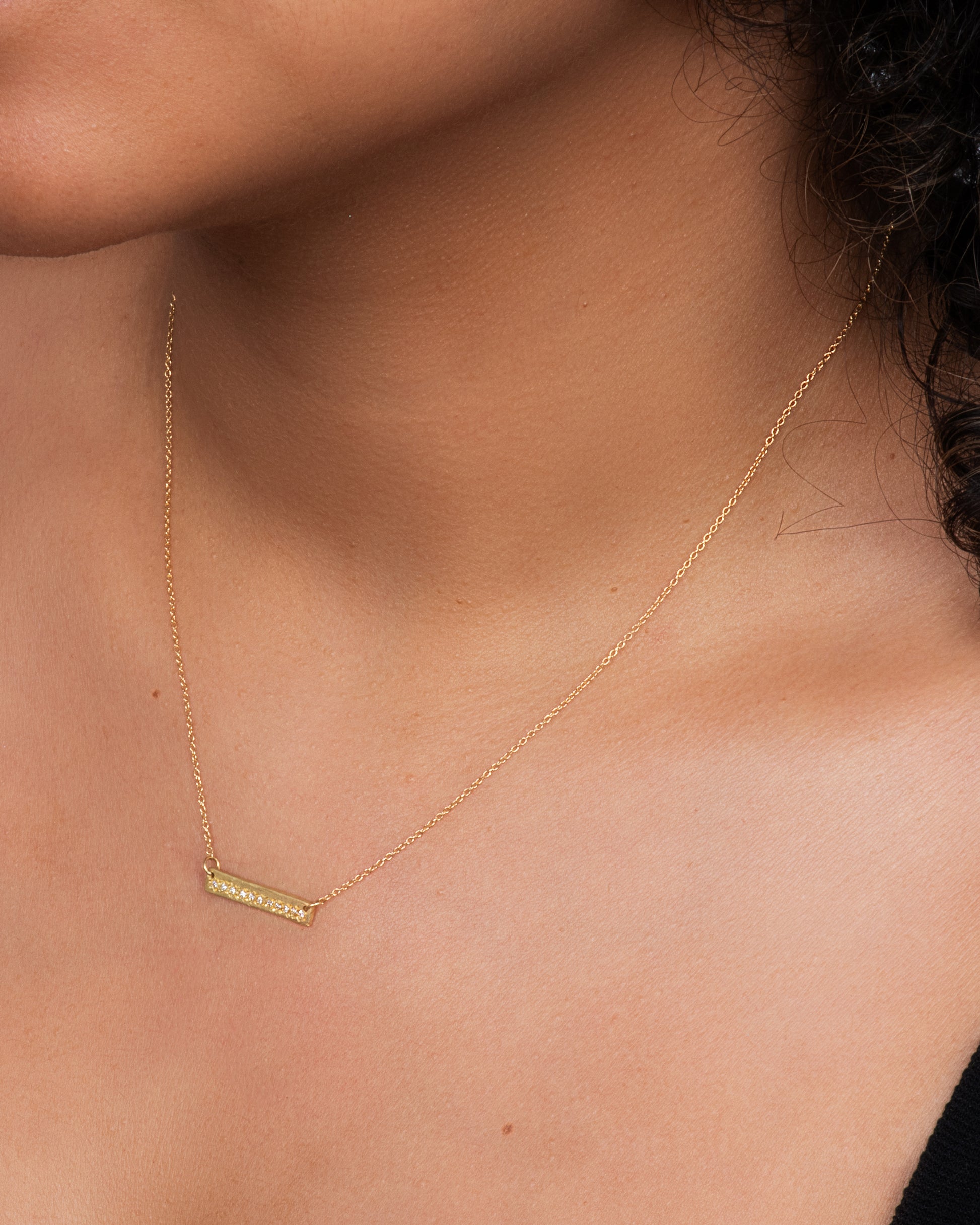 A yellow gold bar necklace with a single row of white diamonds. View on the entire necklace, laying flat. Shown on a neck.