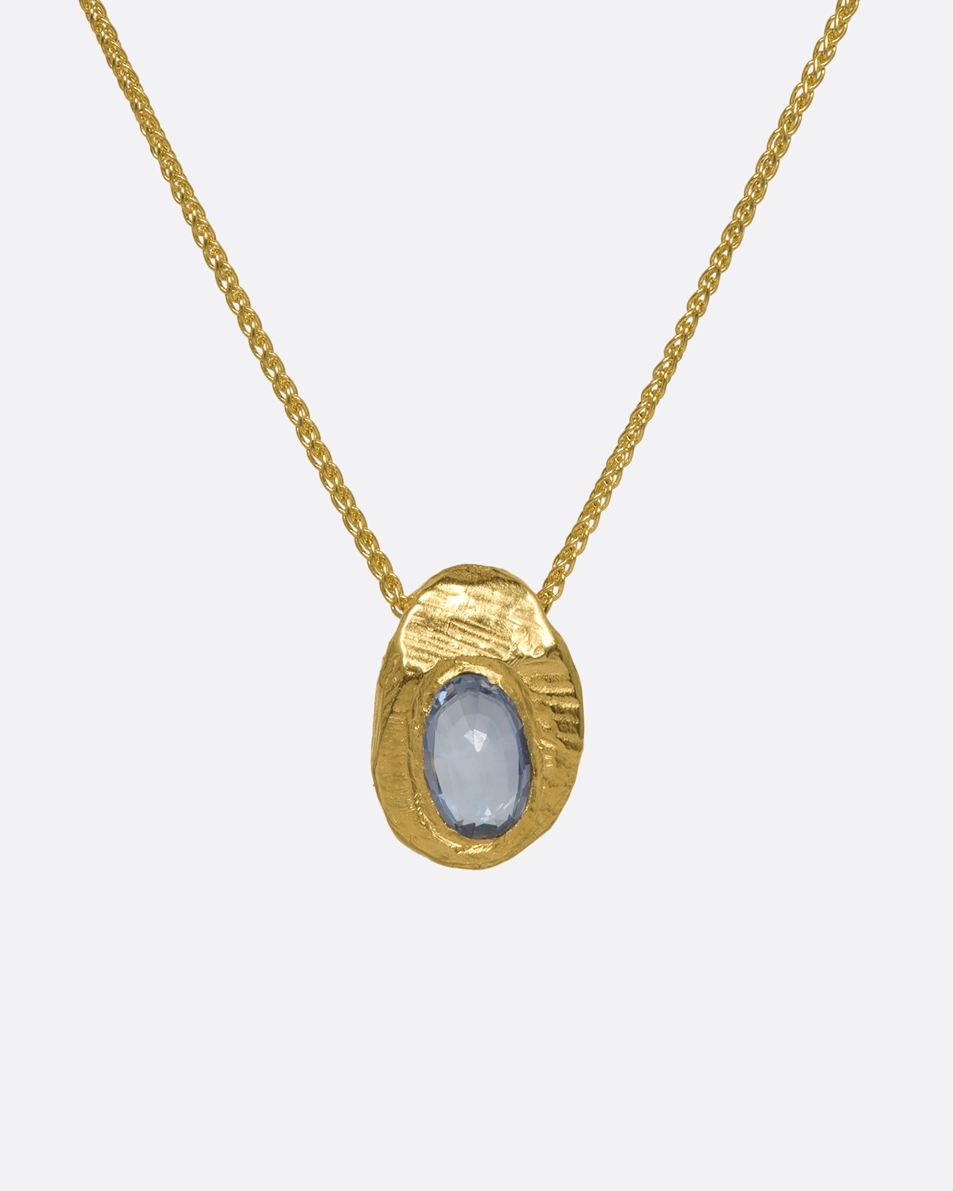 A hand carved textured yellow gold pendant holds a cut sapphire solitaire hanging from a yellow gold chain. View up close from the front.