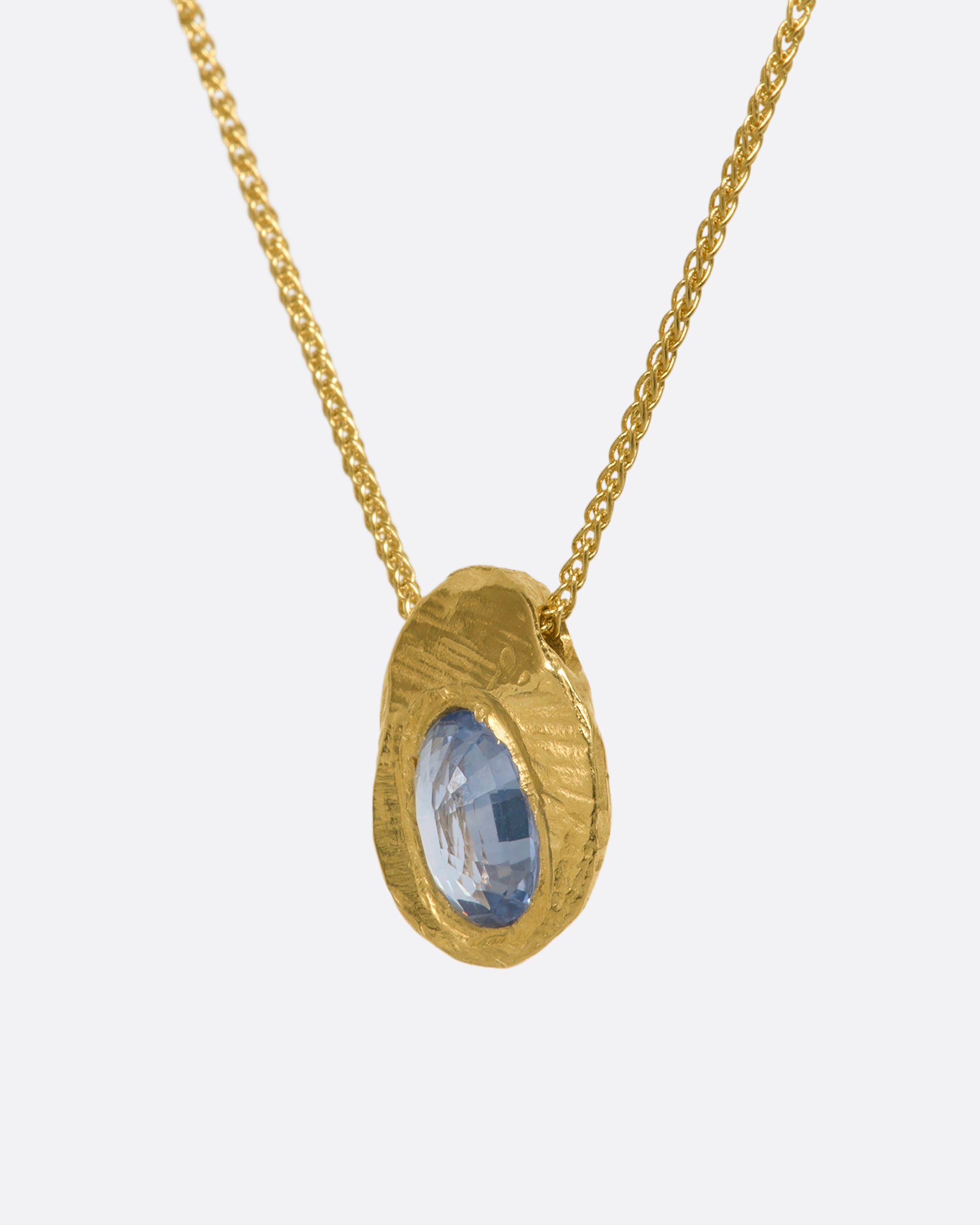 A hand carved textured yellow gold pendant holds a cut sapphire solitaire hanging from a yellow gold chain. View up close from the side.