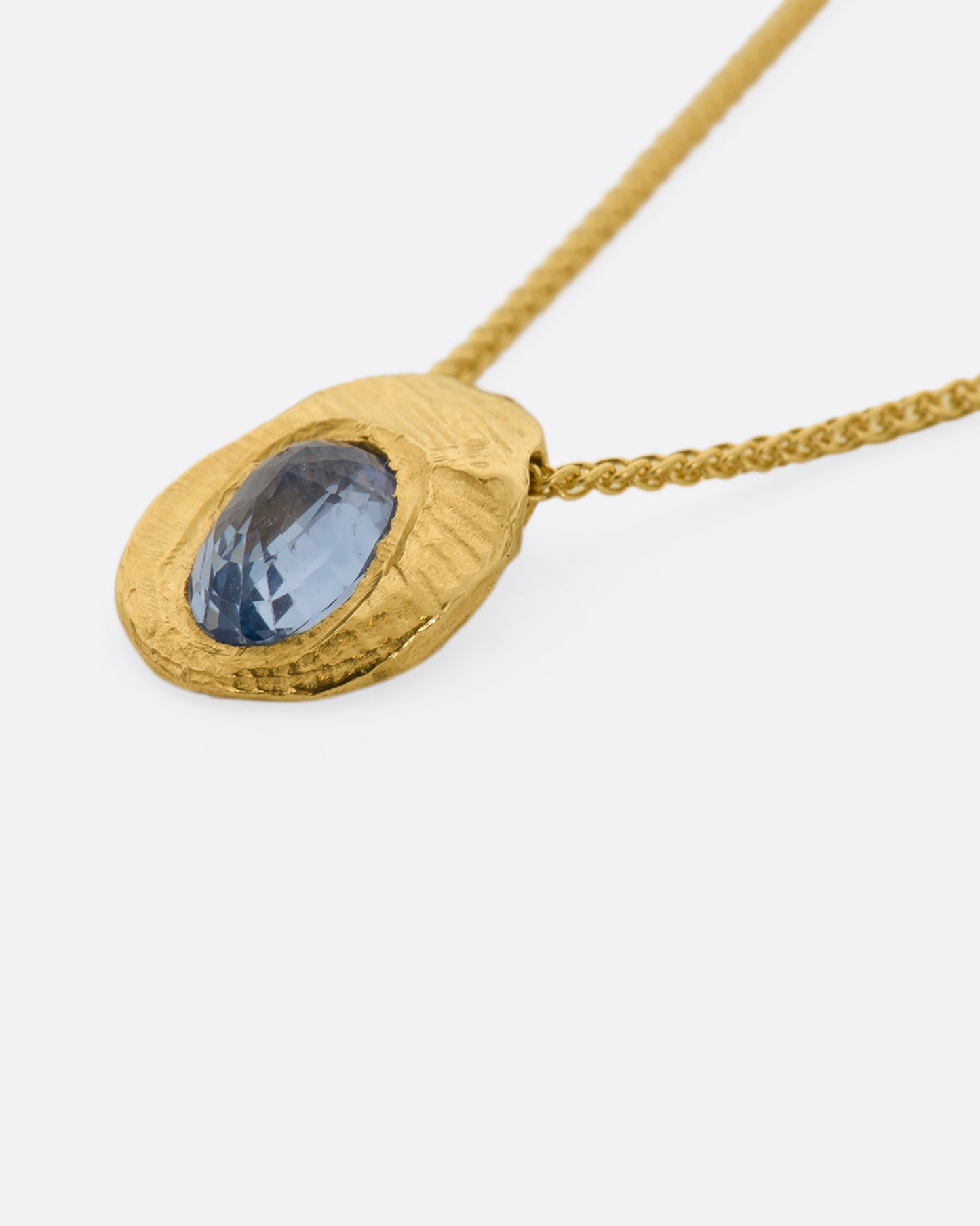 A hand carved textured yellow gold pendant holds a cut sapphire solitaire hanging from a yellow gold chain. View up close laying flat.