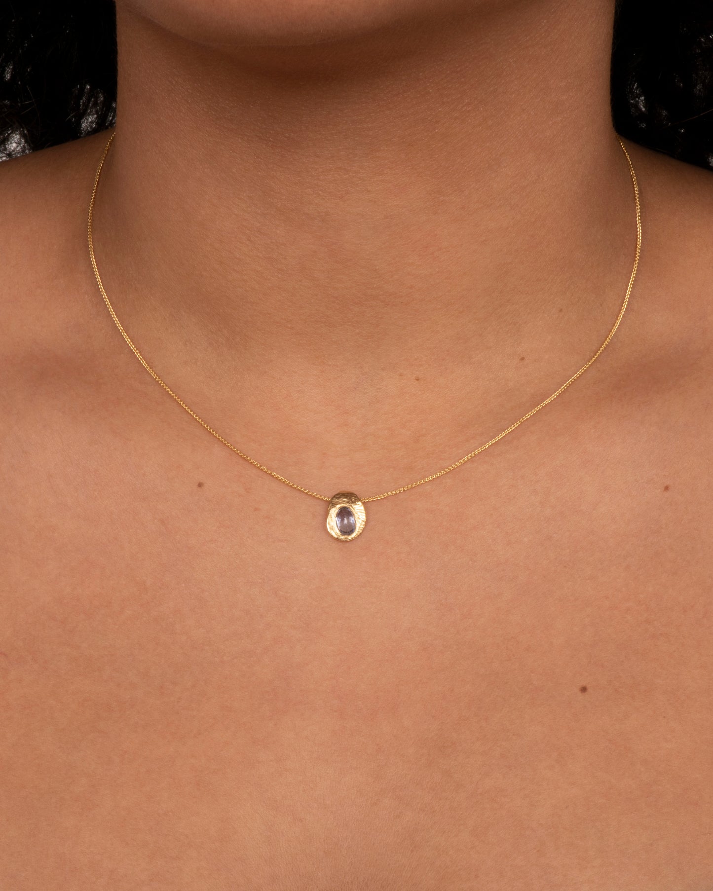 A hand carved textured yellow gold pendant holds a cut sapphire solitaire hanging from a yellow gold chain. View on a neck.