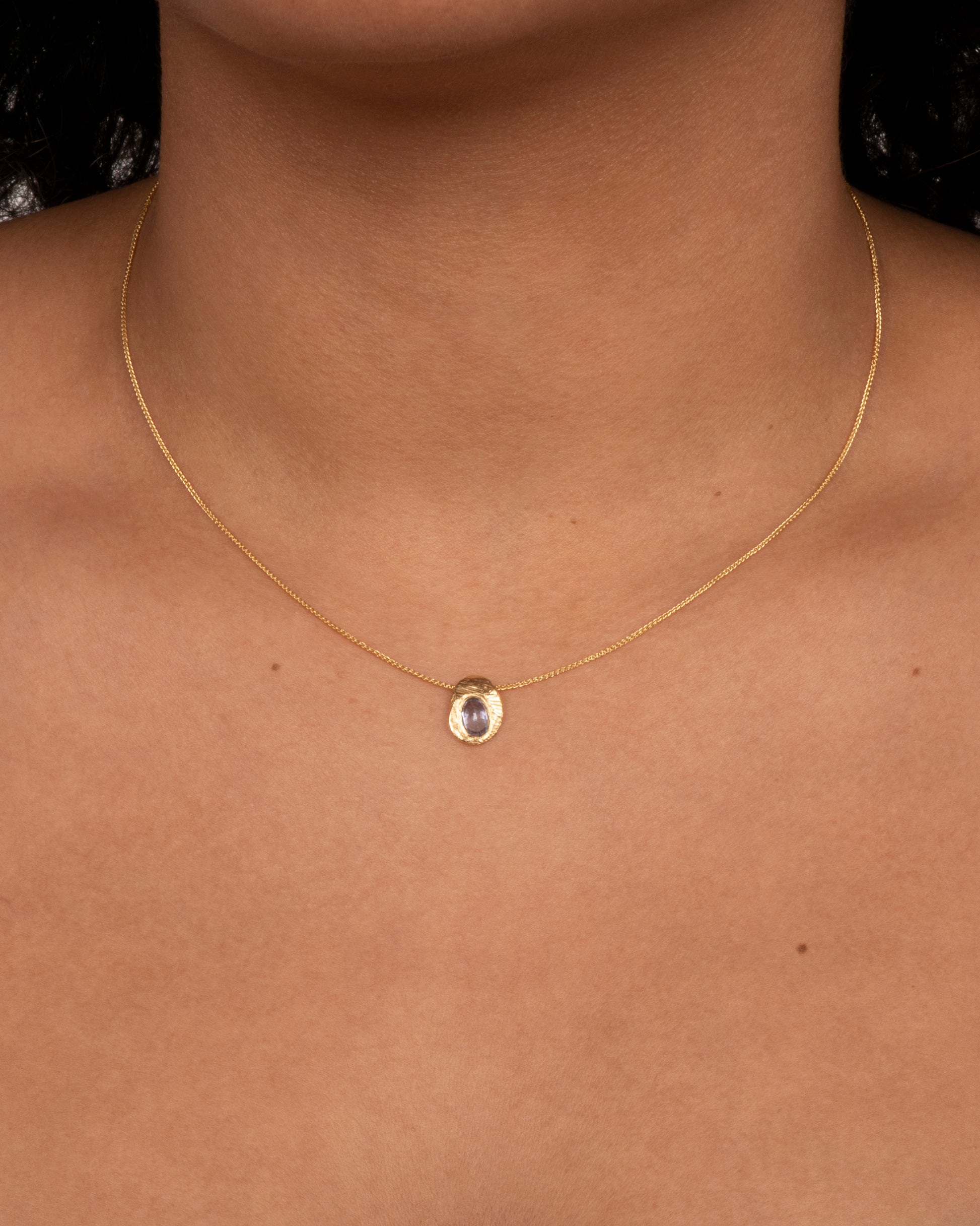 A hand carved textured yellow gold pendant holds a cut sapphire solitaire hanging from a yellow gold chain. View on a neck.