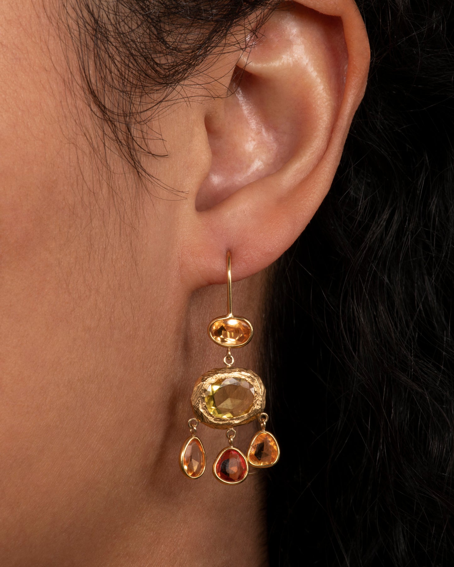 Yellow gold chandelier earrings with three orange sapphires dangling from the bottom. View on an ear.