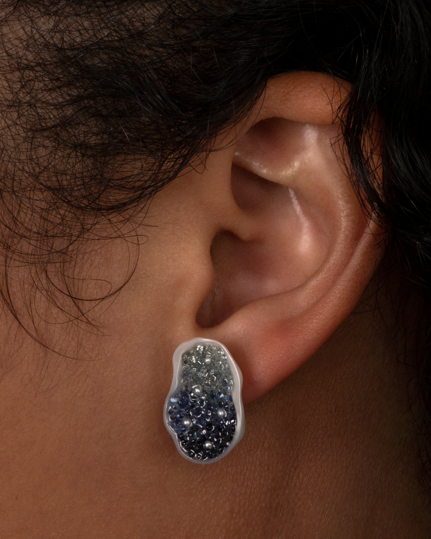 A pair of stud earrings made from halves on one freshwater soufflé pearl, lined with blue sapphires and seed pearls. Shown on ear.