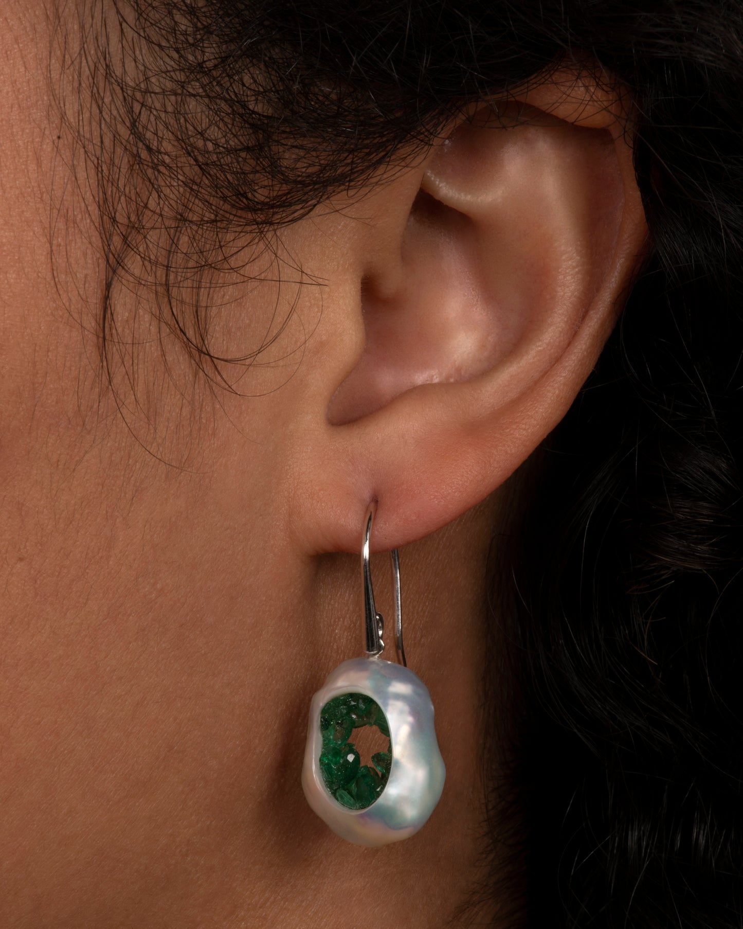 two dangle hook earrings in white gold hanging and viewed from the front. the dangle part is a pearl that has had a hole carved out in the middle and then lined with emeralds, to create a geode like appearance. View on an ear.