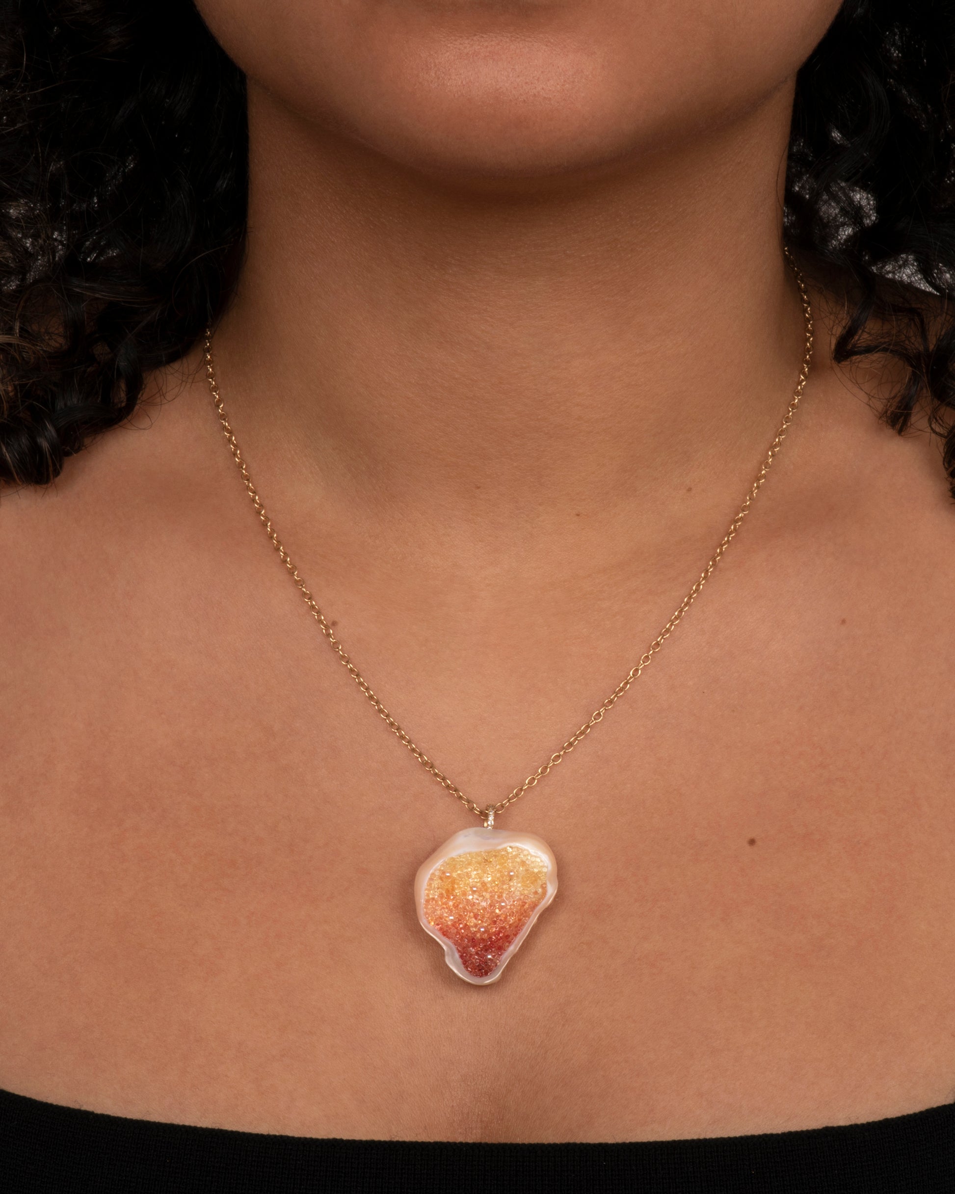 A yellow gold necklace with a soufflé pearl pendant lined with gradient orange sapphires and a pave diamond bail. View on a neck.