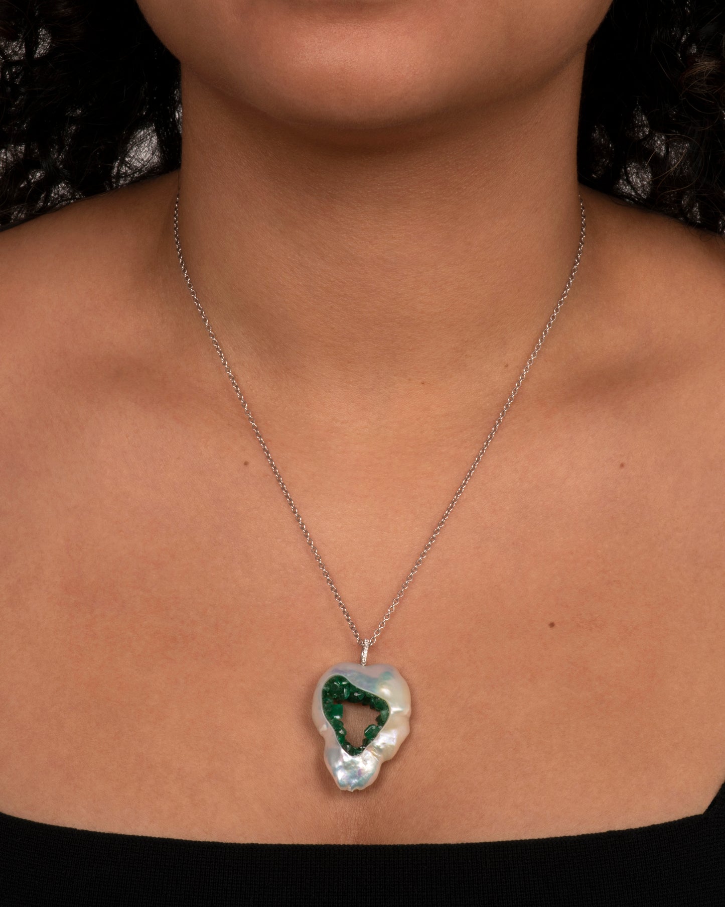 White gold necklace with a souffle pearl pendant lined in emeralds and a pave bale. View on a neck.