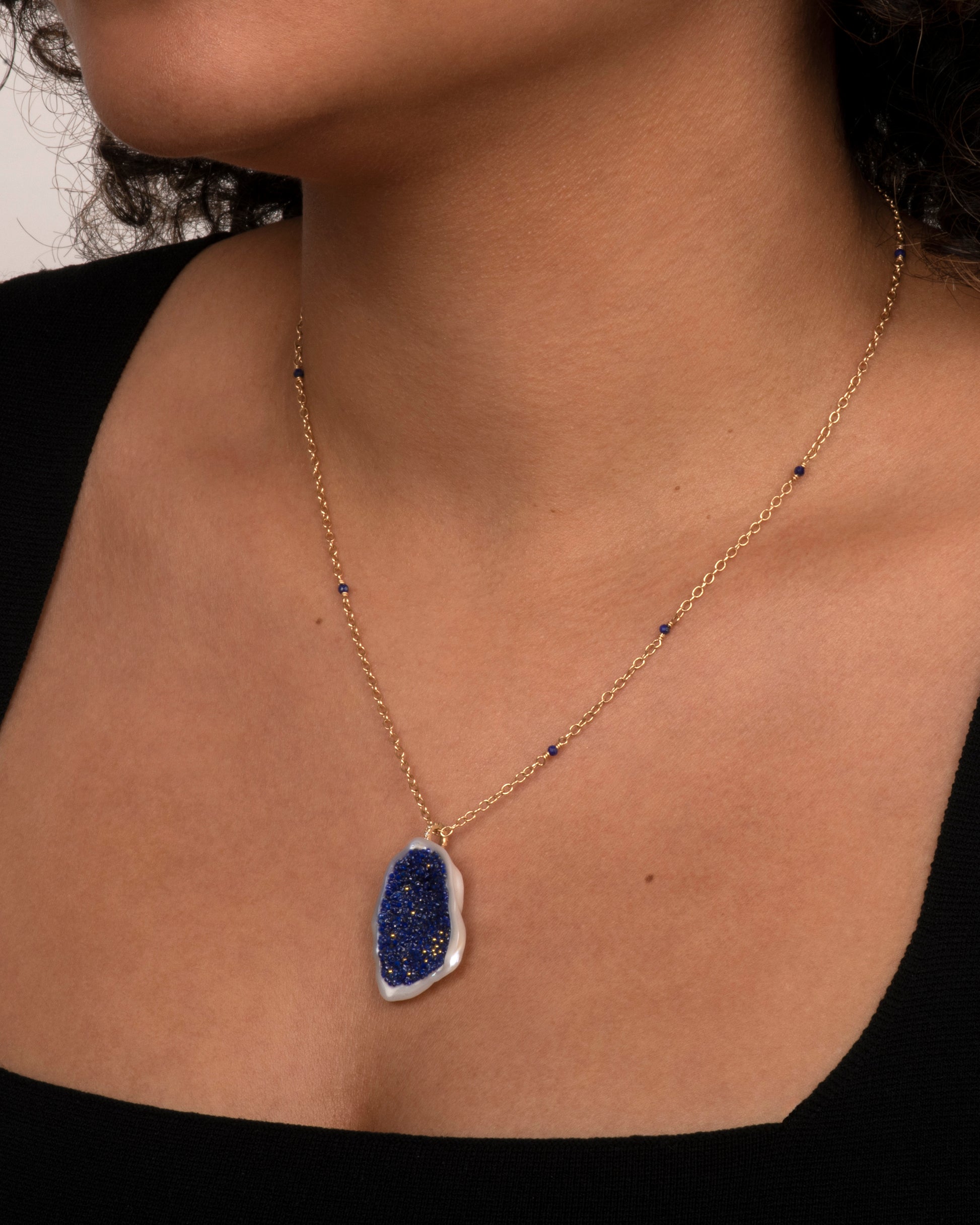 A yellow gold necklace with a hollowed out pearl pendant filled with lapis and 22k gold beads. Shown worn on the neck.