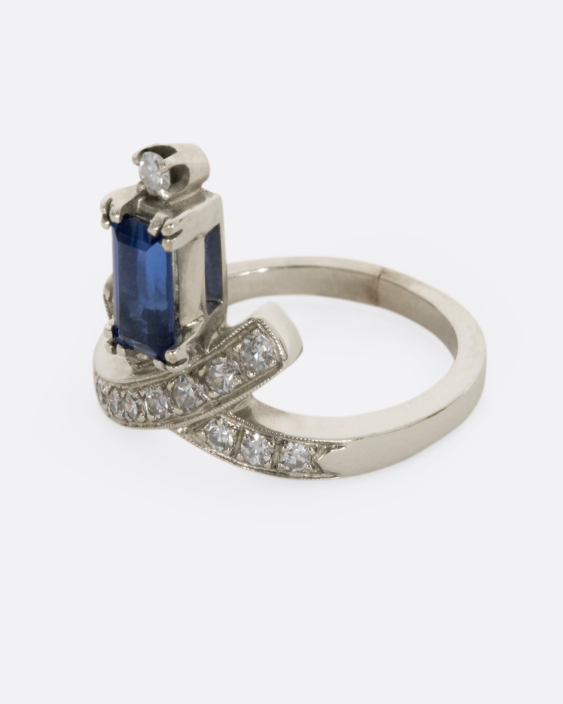 A white gold crossed band ring with a baguette blue sapphire topped with a diamond. View from the side.