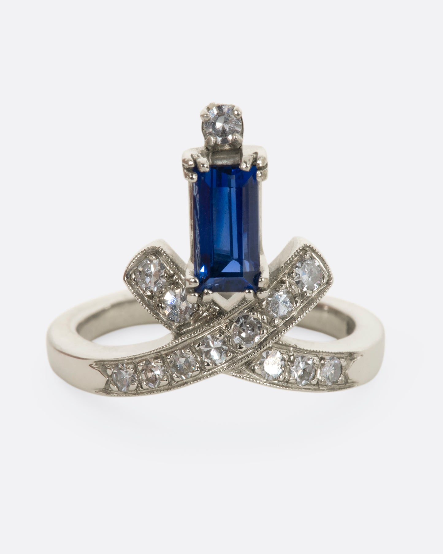 A white gold crossed band ring with a baguette blue sapphire topped with a diamond. View from the front.